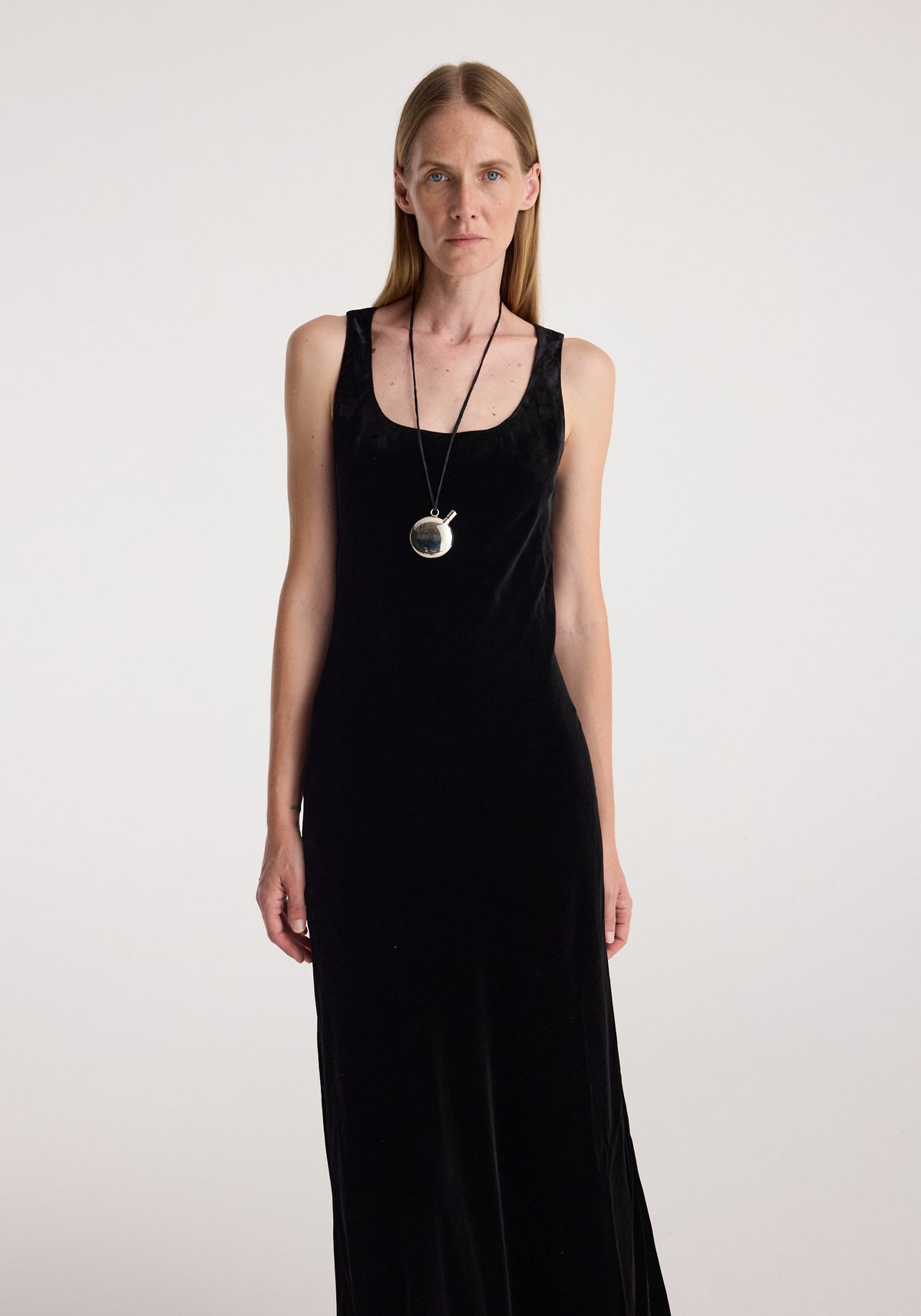 Woman wearing a long black scoop-neck velvet dress | black with a sleeveless design and a pendant necklace against a plain light background.