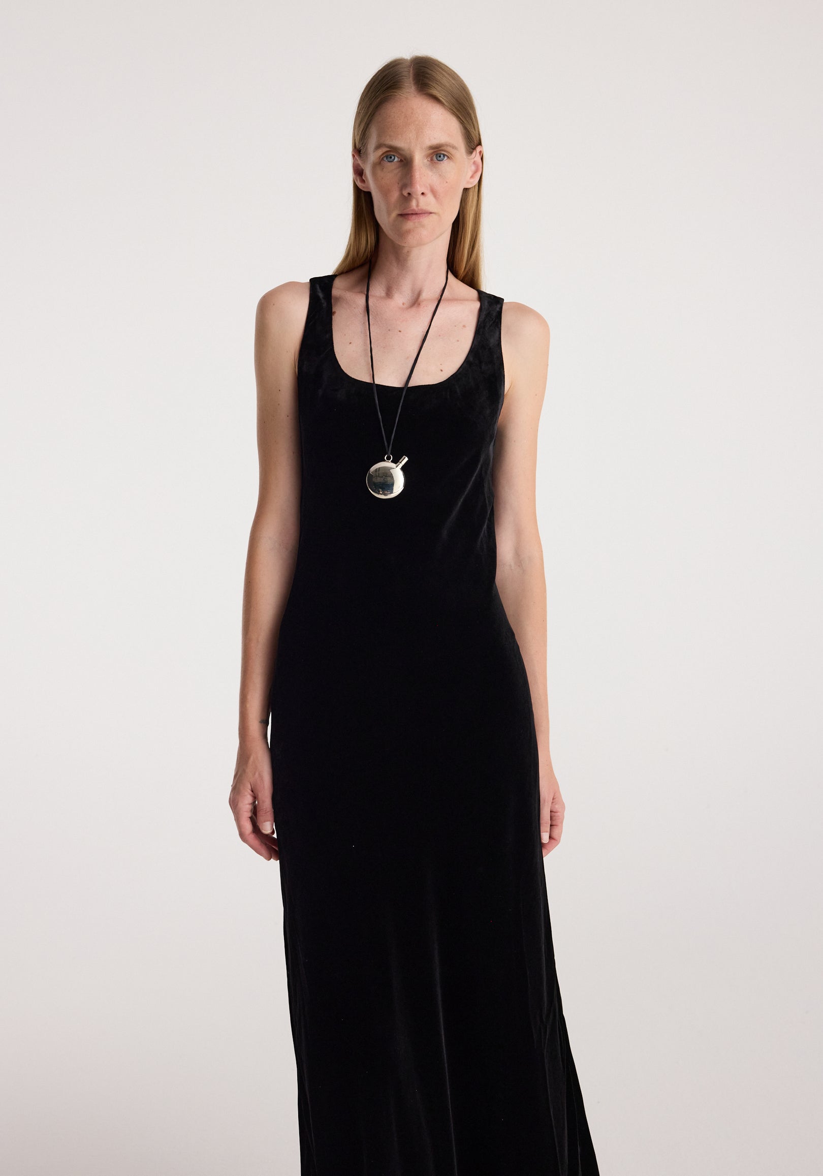 Woman wearing a long black scoop-neck velvet dress | black with a sleeveless design and a pendant necklace against a plain light background.