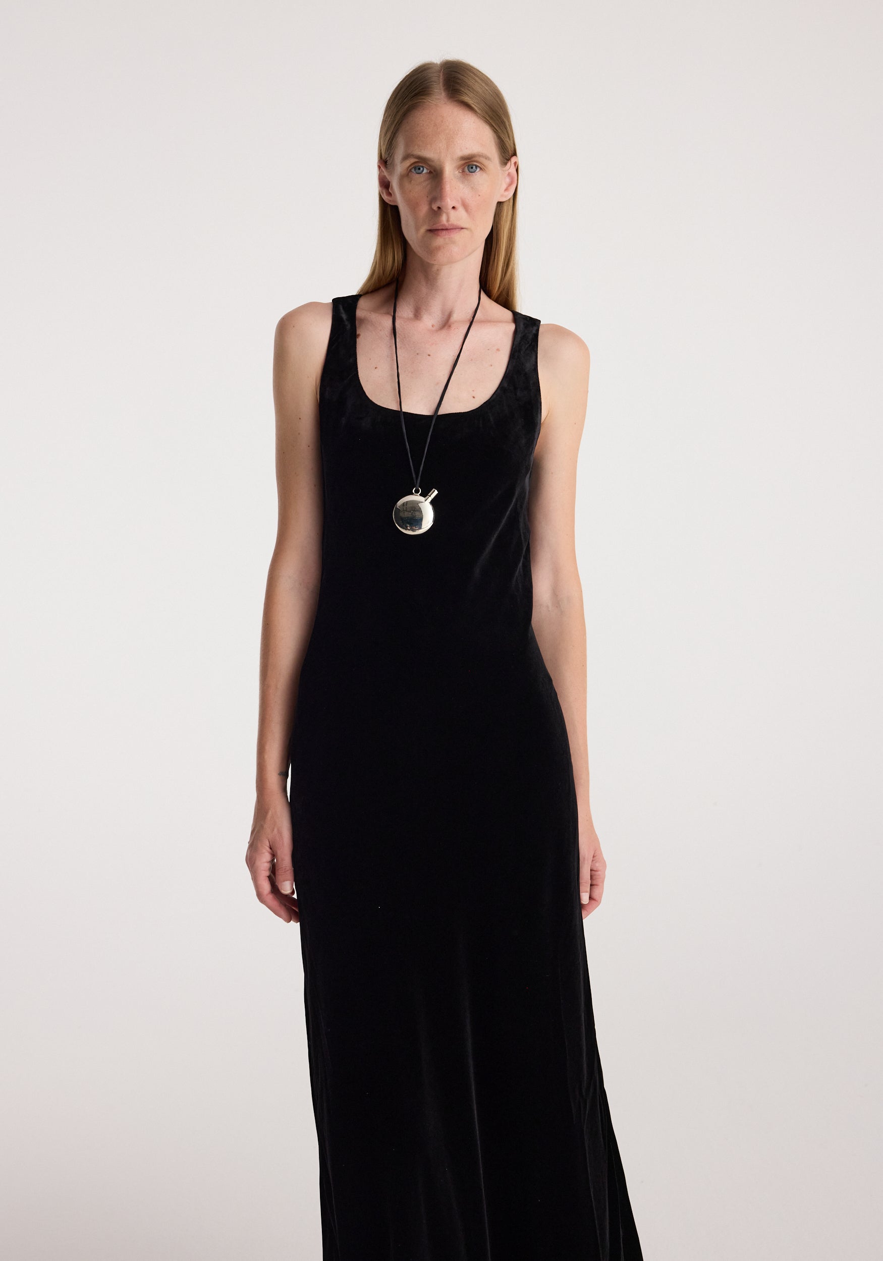 Woman wearing a long black dress and a Silver perfume bottle necklace | silver pendant on a long chain around her neck against a plain white background.