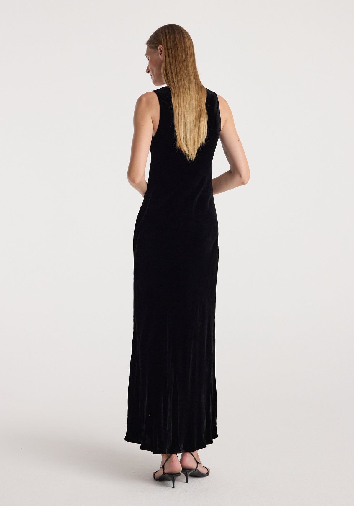 Woman wearing a sleeveless long black scoop-neck velvet dress | black, shown from the back with straight hair and black heeled sandals.