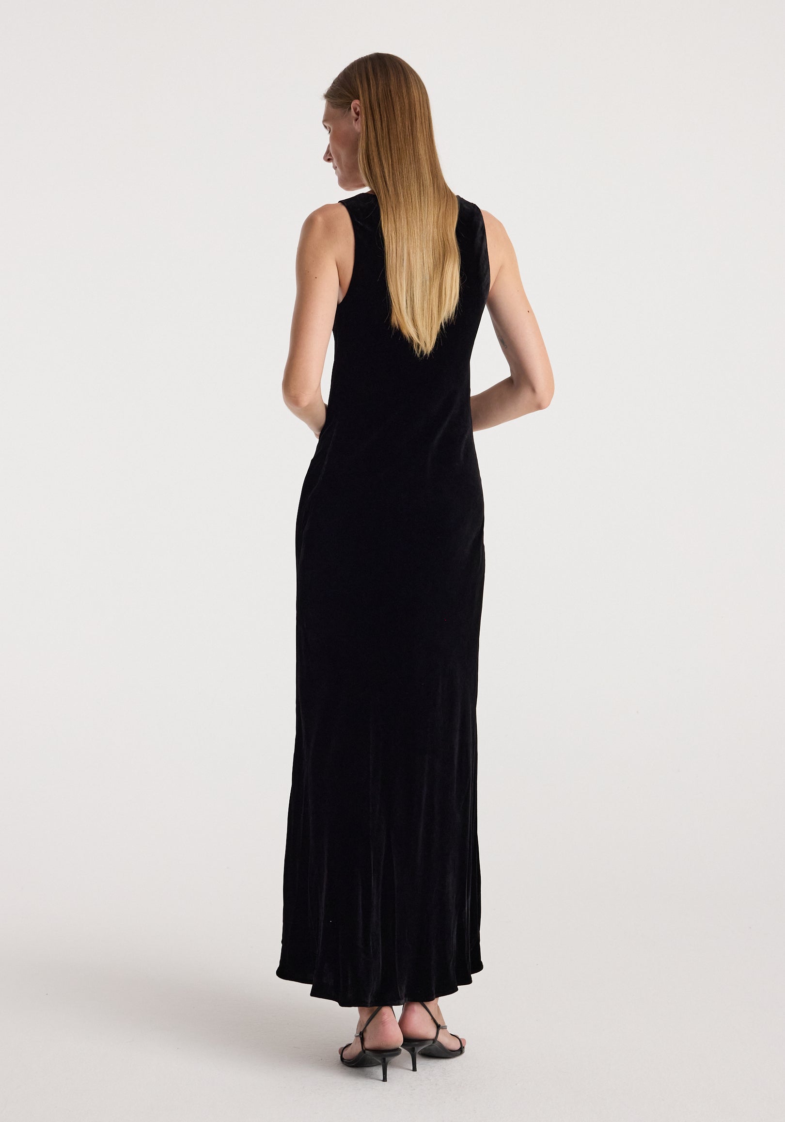 Woman wearing a sleeveless long black scoop-neck velvet dress | black, shown from the back with straight hair and black heeled sandals.