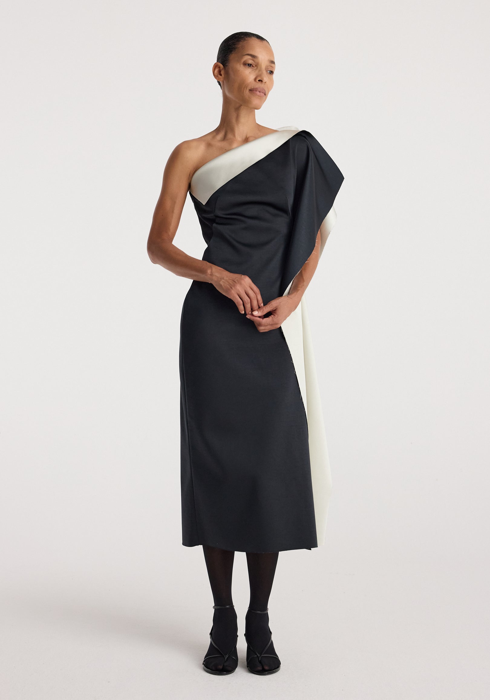 Woman wearing a sculptural double-satin off-shoulder dress | black/white with a sleek, elegant design and draped asymmetrical neckline, posing with arms crossed