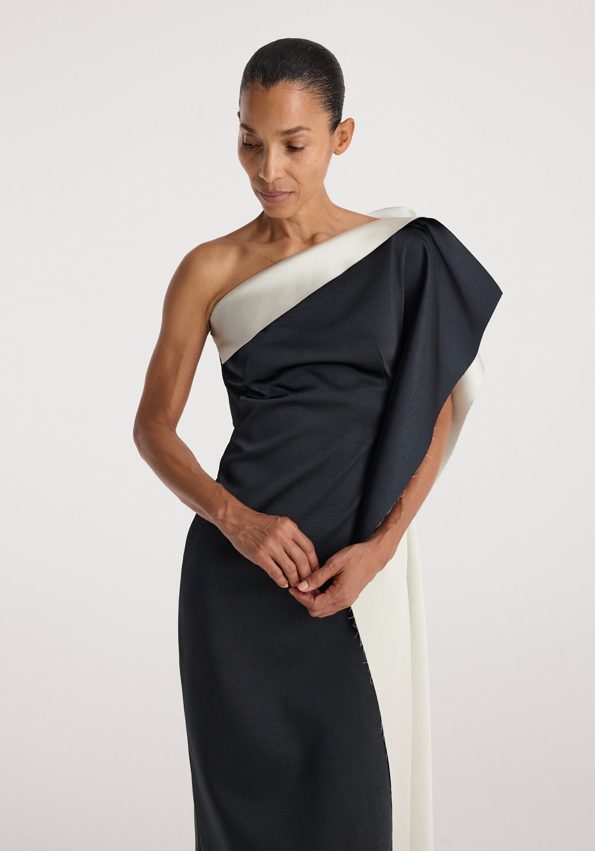 Woman wearing sculptural double-satin off-shoulder dress | black/white with an asymmetrical neckline and draped design against a plain light background.