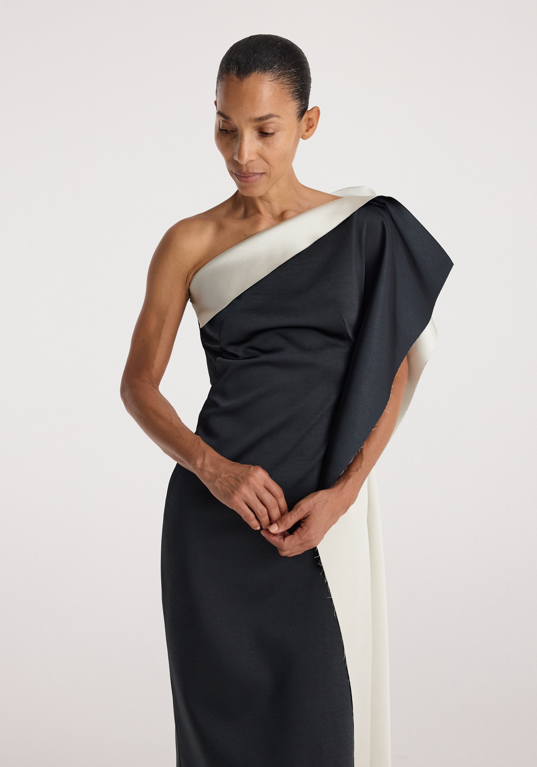 Woman wearing sculptural double-satin off-shoulder dress | black/white with an asymmetrical neckline and draped design against a plain light background.