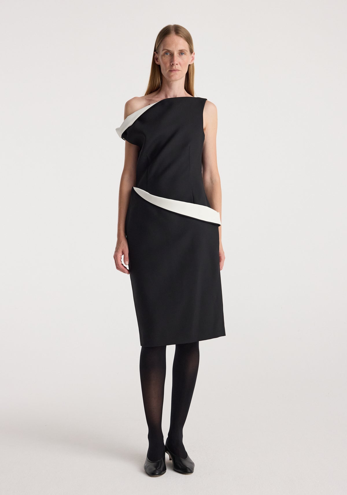 Woman wearing a sculptural off-shoulder dress | black with white sculptural accents along the neckline and waist, paired with black tights and black heels, standing against a plain background.