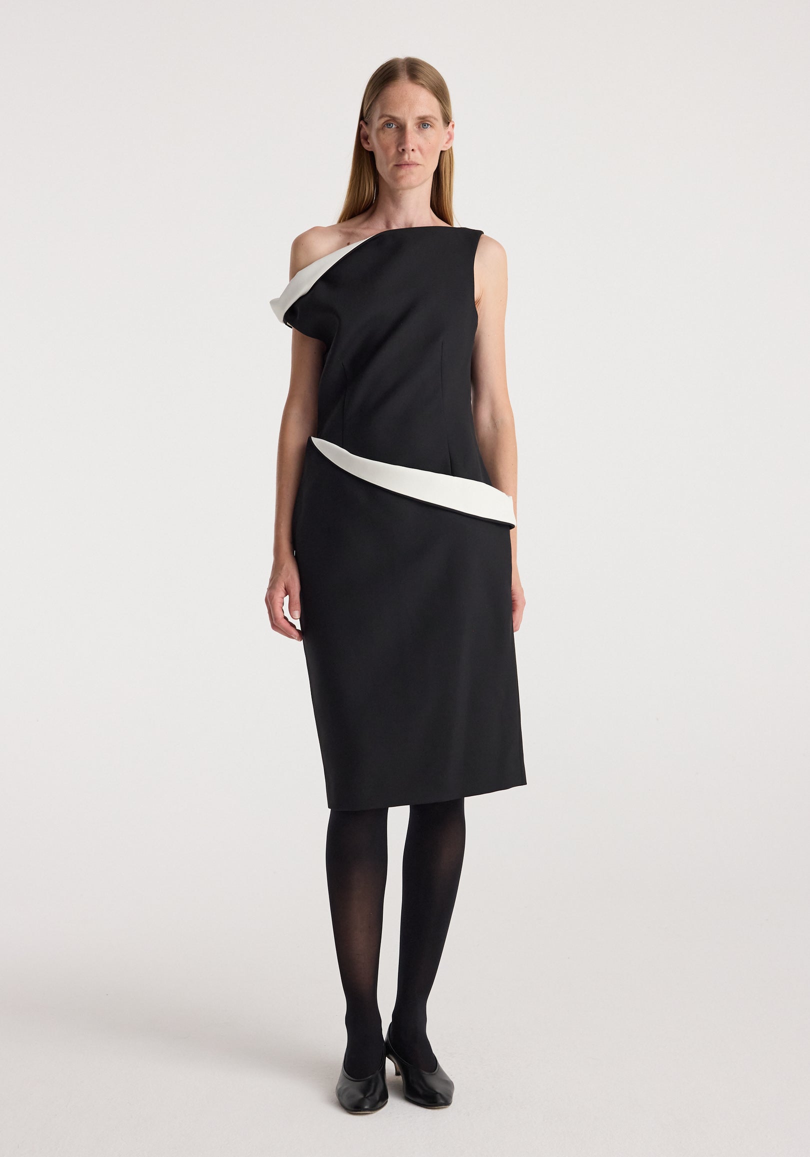 Woman wearing a sculptural off-shoulder dress | black with white sculptural accents along the neckline and waist, paired with black tights and black heels, standing against a plain background.