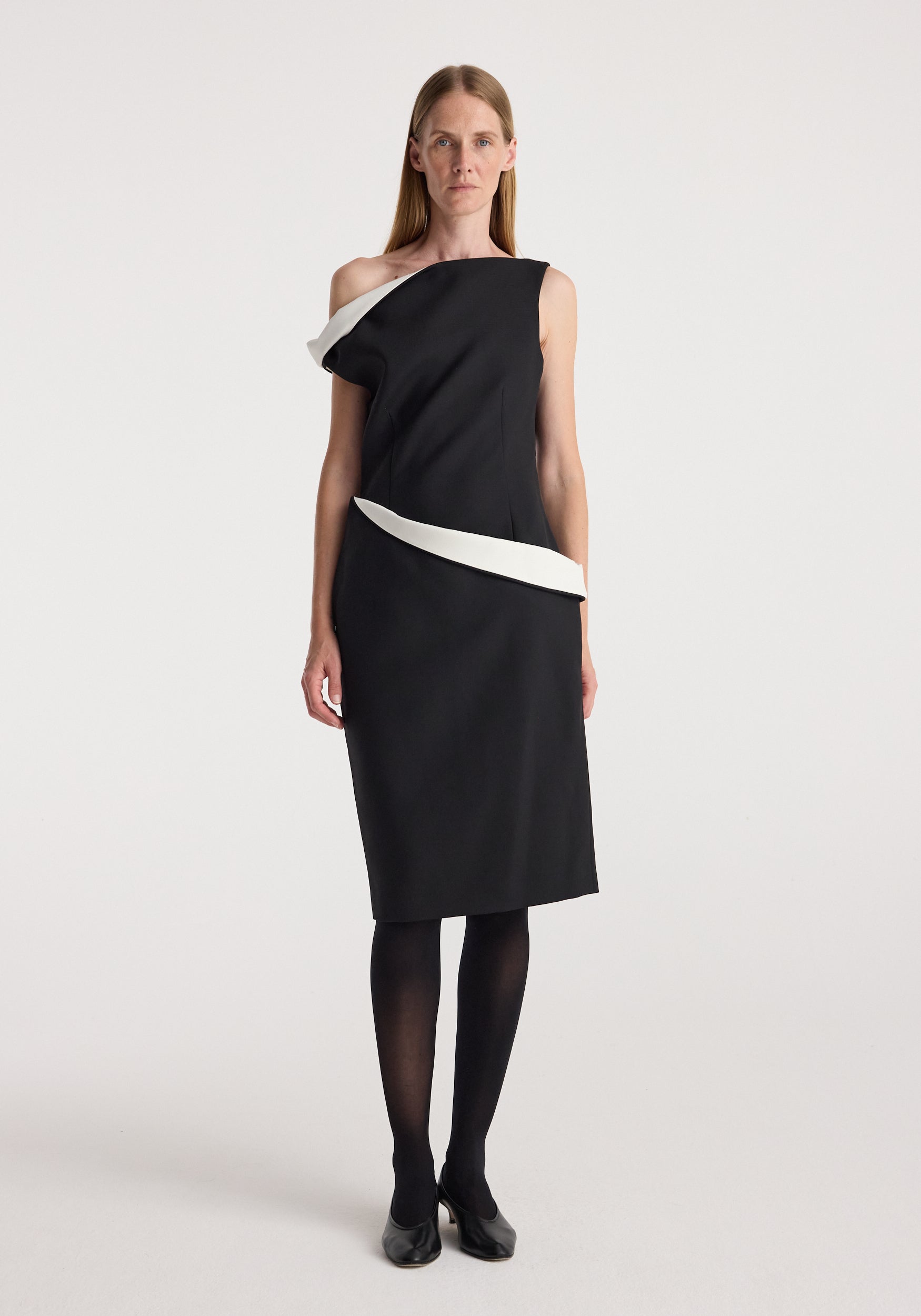 Woman wearing a sculptural off-shoulder dress | black with white sculptural accents along the neckline and waist, paired with black tights and black heels, standing against a plain background.