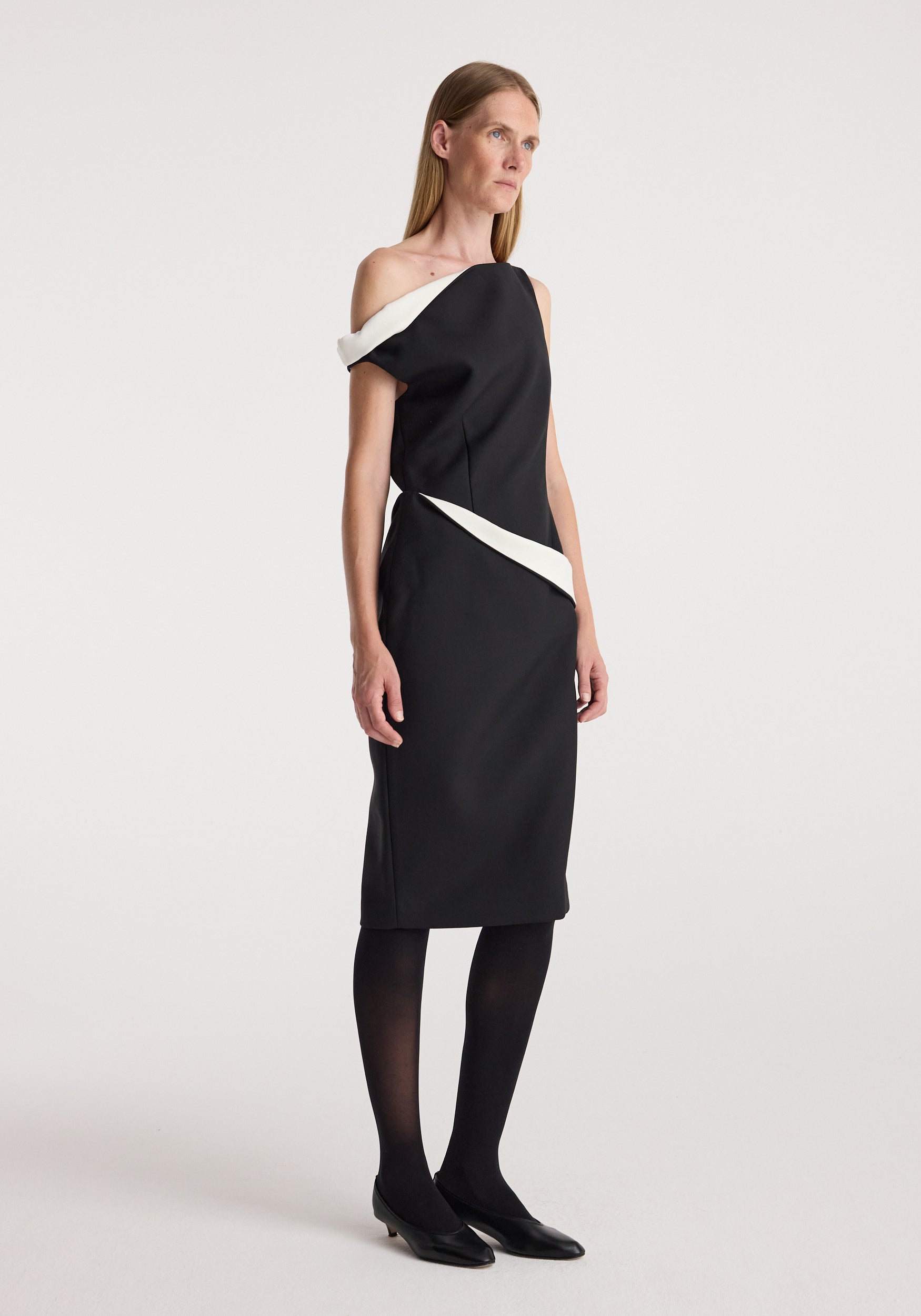 Woman wearing a sculptural off-shoulder dress | black with white accents, paired with black tights and low black heels, standing against a plain background.