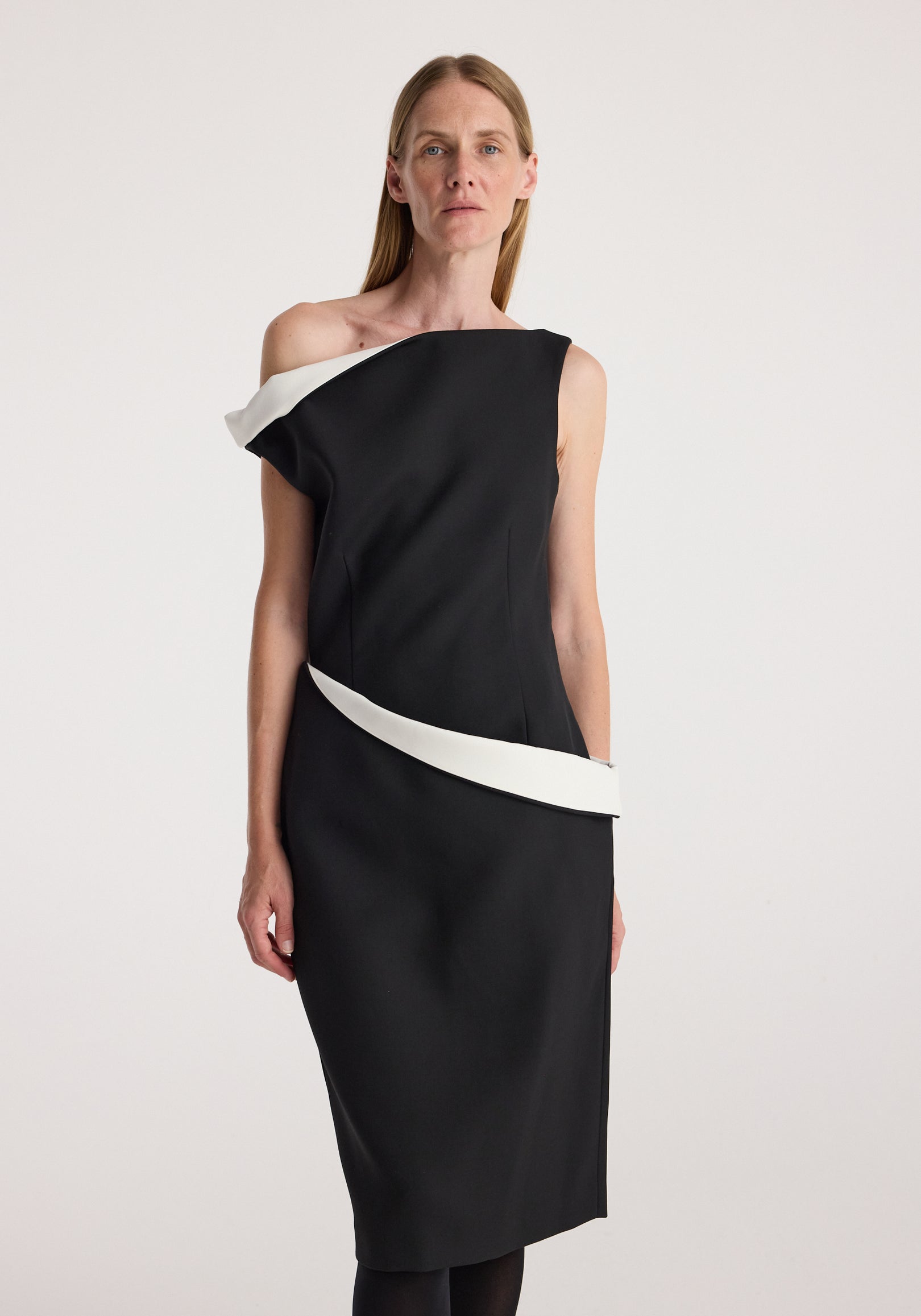 Woman wearing a sculptural off-shoulder dress | black with a white accent detail along the neckline and waist, standing against a plain light background.