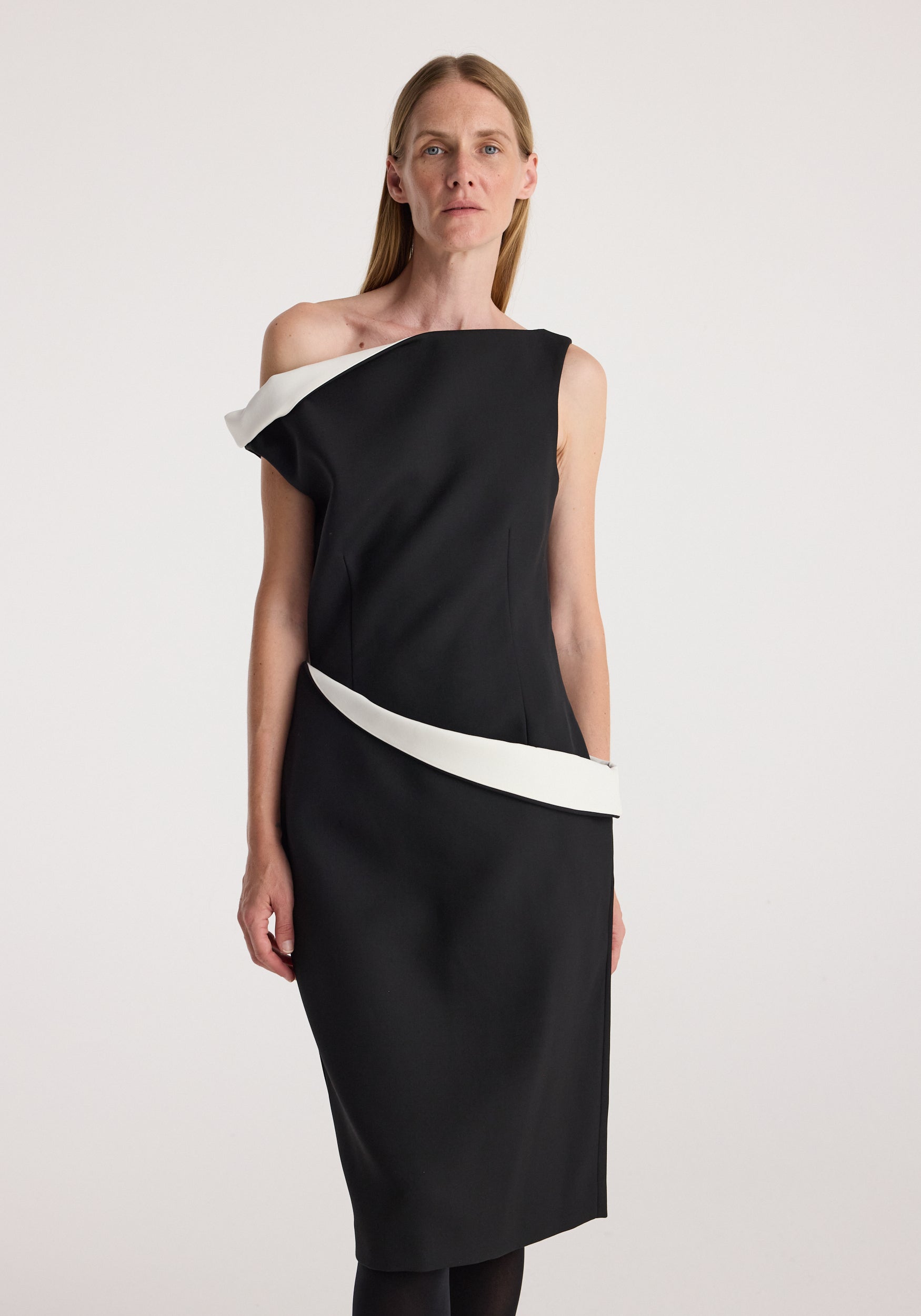 Woman wearing a sculptural off-shoulder dress | black with a white accent detail along the neckline and waist, standing against a plain light background.