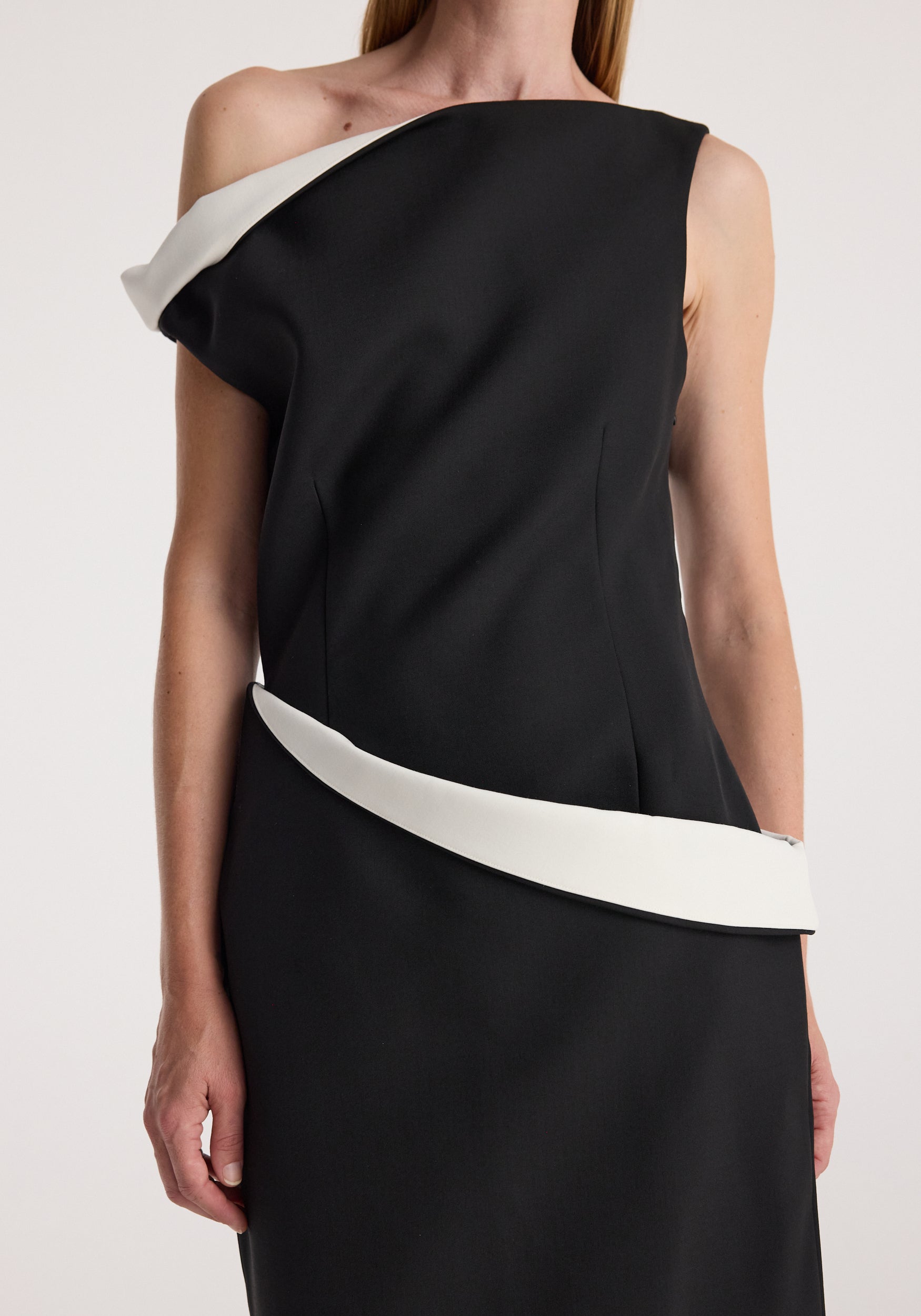 Woman wearing a sculptural off-shoulder dress | black featuring asymmetrical white trim along the neckline and waist against a neutral background.