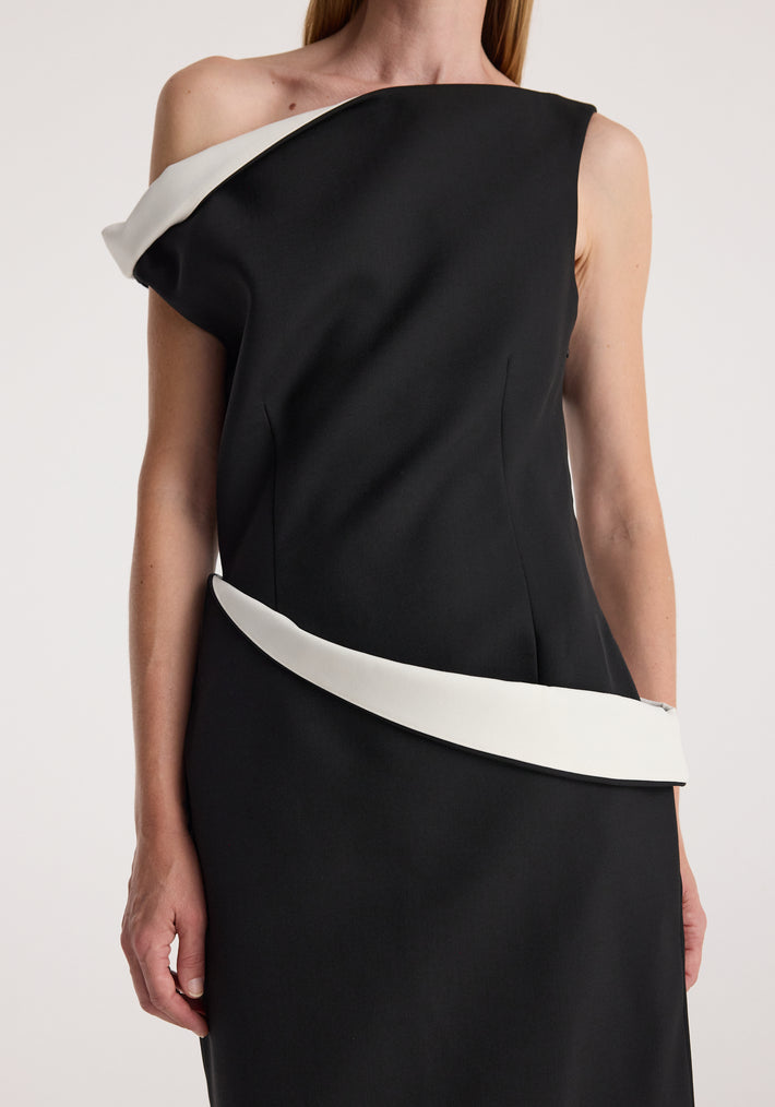 Woman wearing a sculptural off-shoulder dress | black featuring asymmetrical white trim along the neckline and waist against a neutral background.