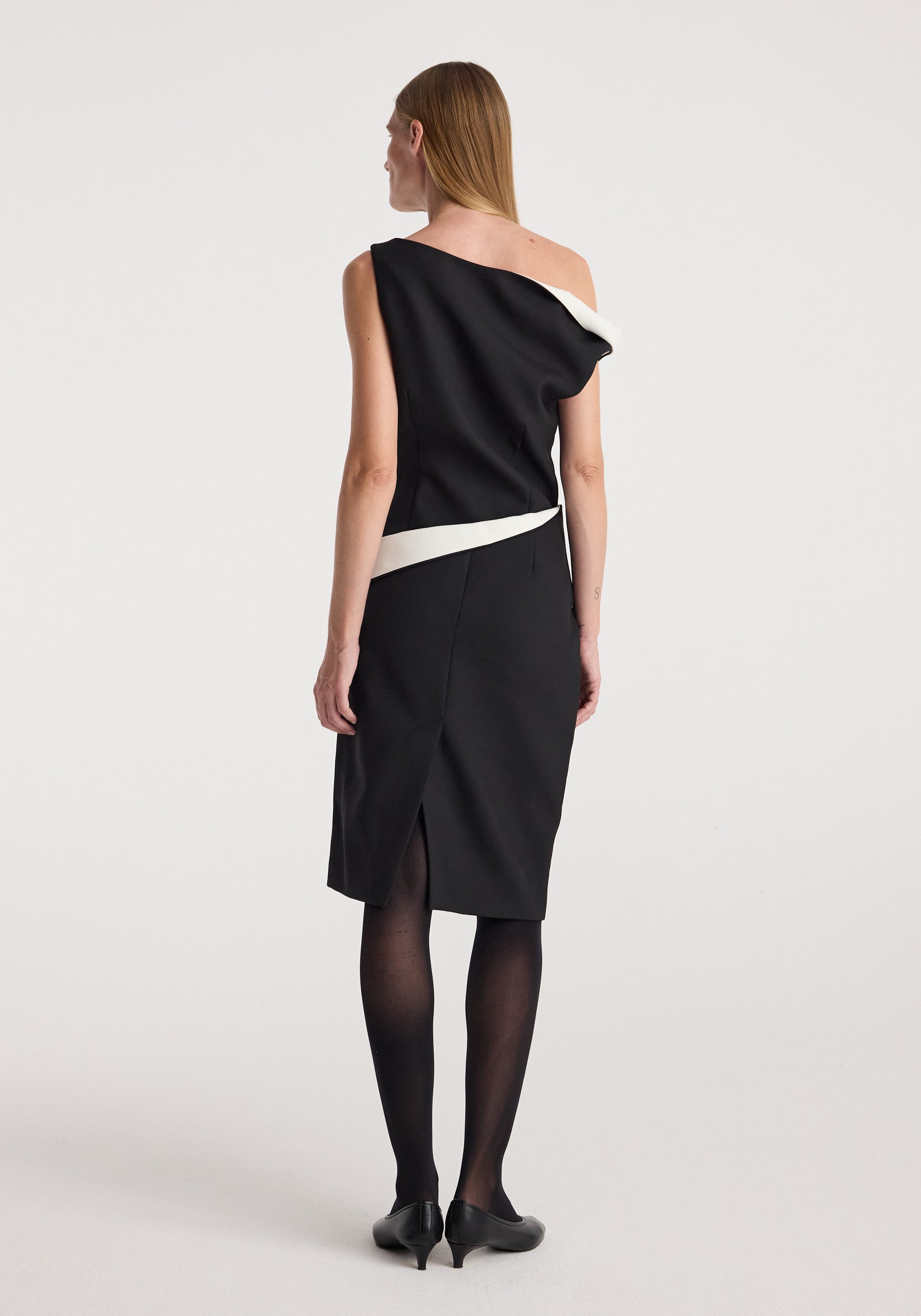 Woman wearing a Sculptural off-shoulder dress | black with a contrasting white accent, standing with her back to the camera against a plain background.