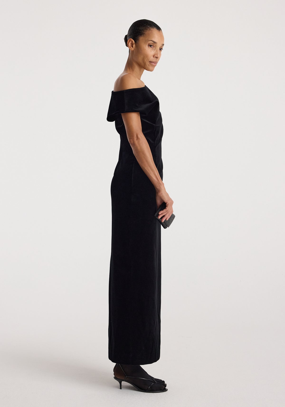 Sculptural off-shoulder velvet gown | black worn by a model in side profile, featuring a long fitted silhouette and elegant draped neckline with black heels