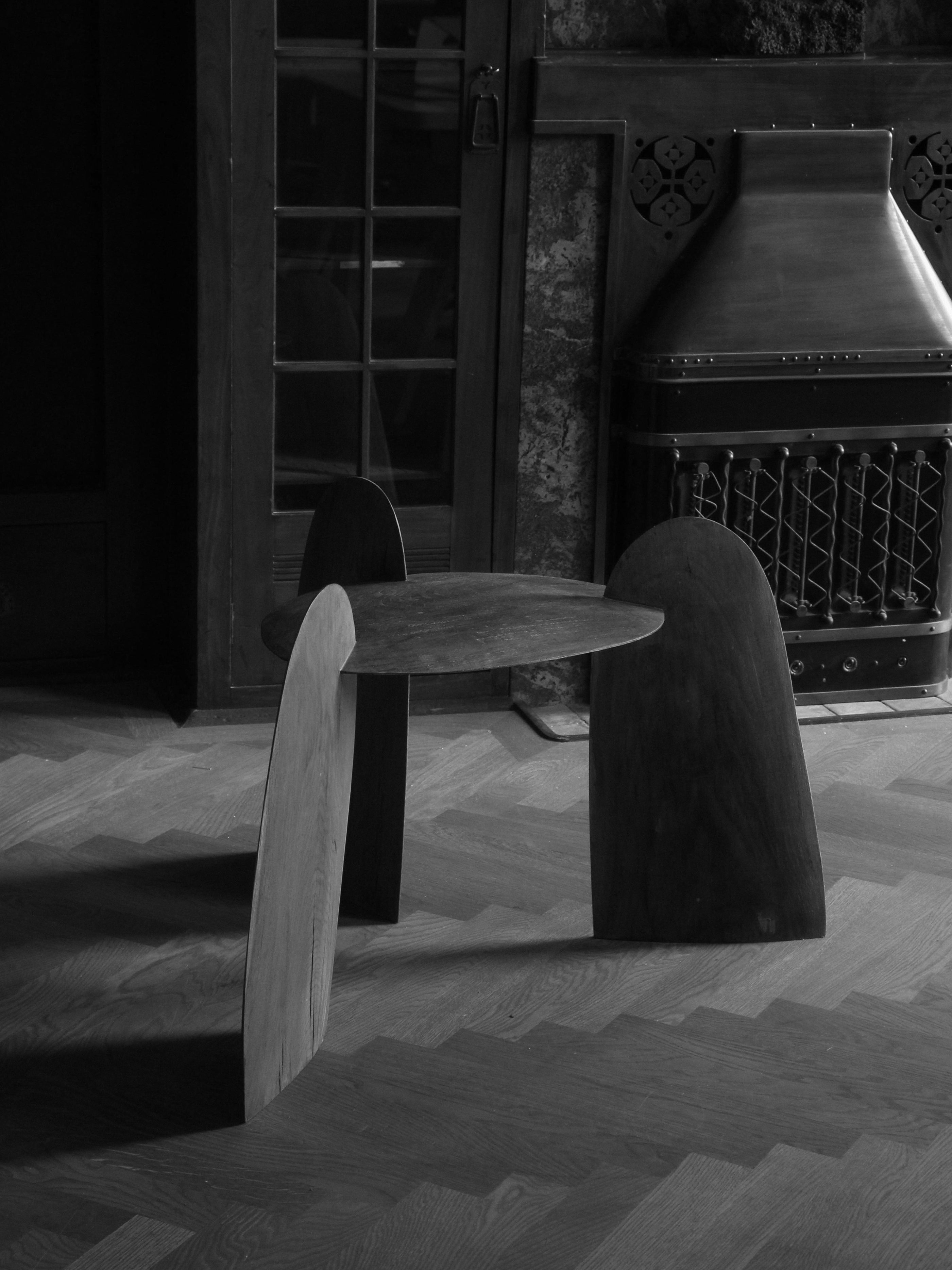 Sculpted Stool | 4673 featuring a unique three-legged design with smooth, curved wooden panels set on a parquet floor near a vintage fireplace and glass-paneled door in a dimly lit room.