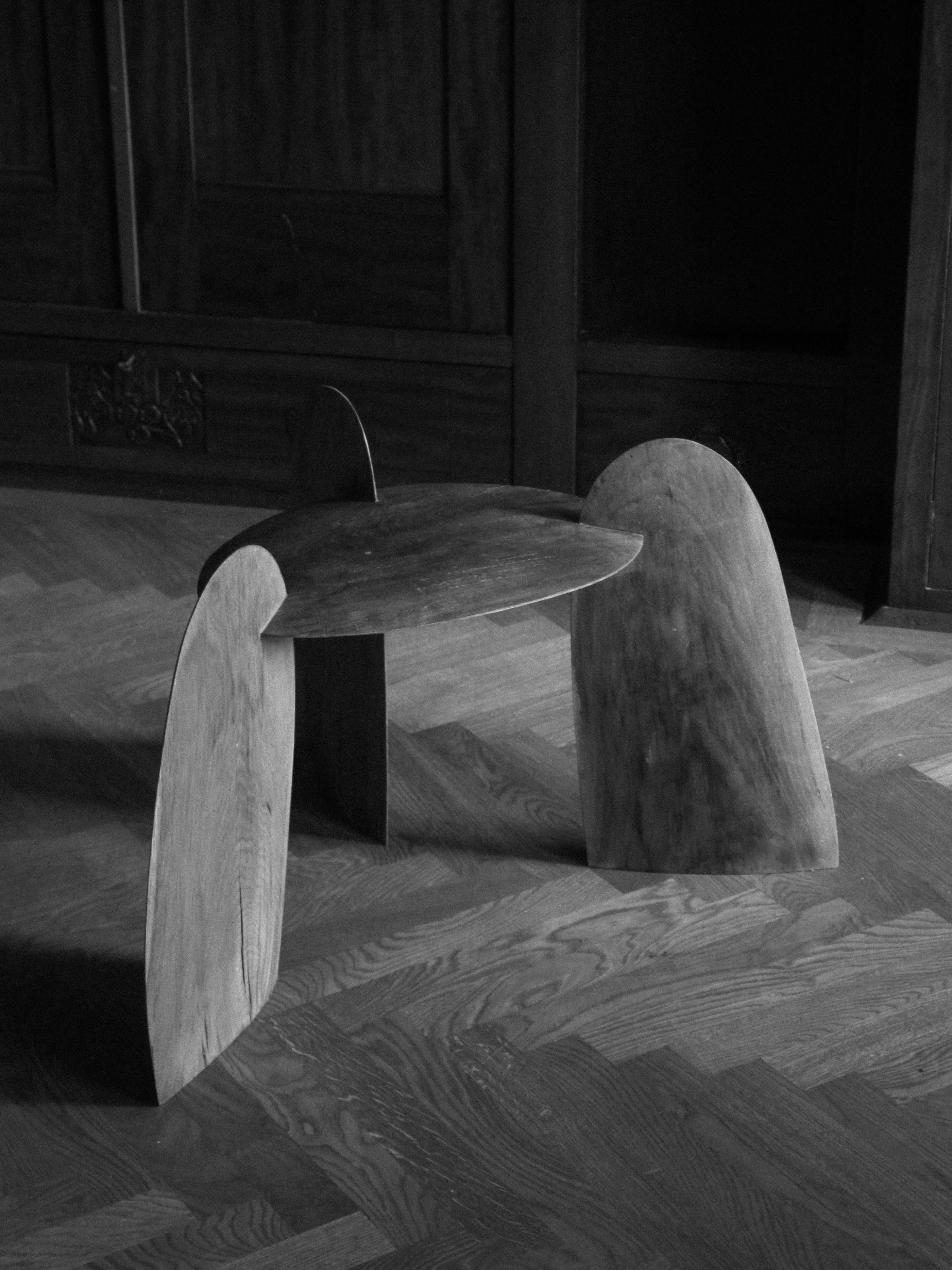 Sculpted Stool | 4673 made of smooth, curved wooden panels forming a minimalist, sculptural three-legged seat on a wooden floor with a dark background.