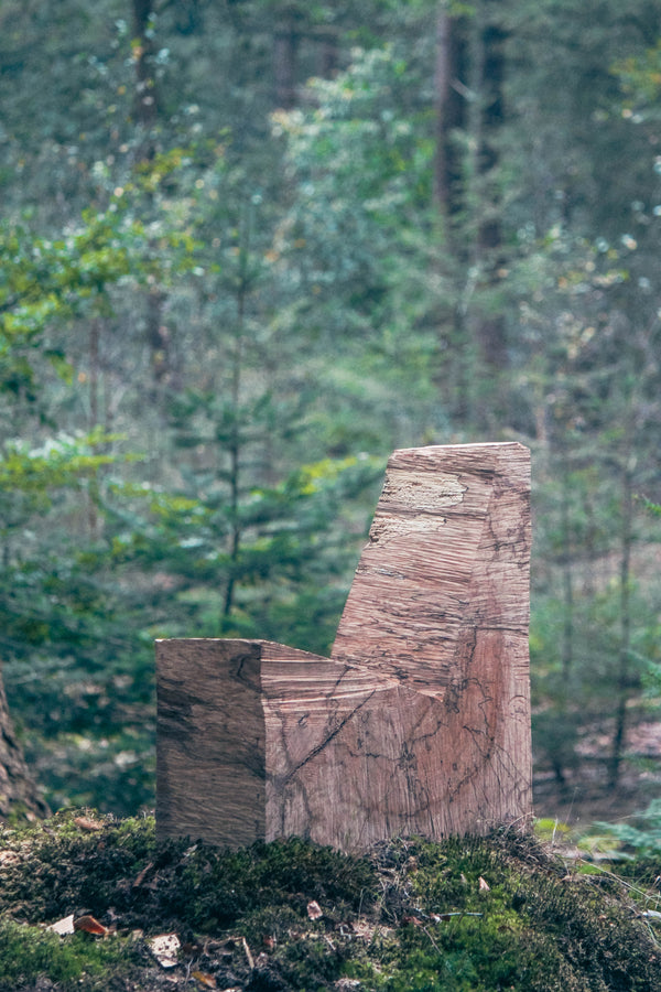 Brutalist Chair | 5788, a minimalist oak block with visible saw cuts, sits on a mossy forest floor, blending sculpture and function.