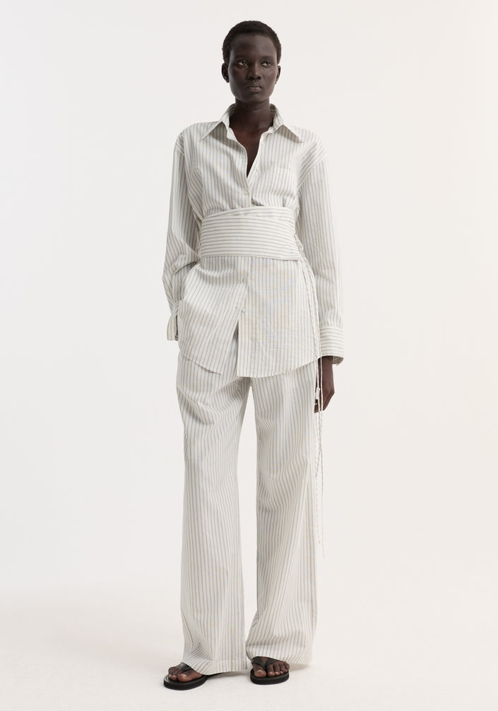 Model wearing a Silk Stripe Belted Shirt with wide sleeves and a wrap belt, paired with matching pin-stripe trousers and black flat sandals.