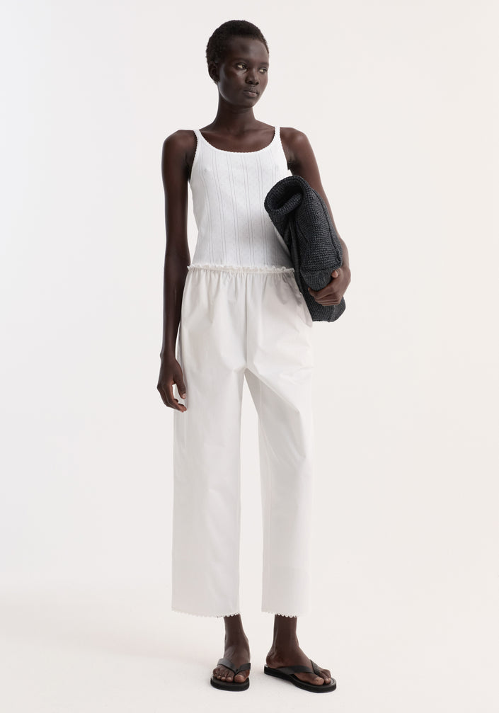 Model in Embroidered poplin trousers, optic white, with scalloped edge, high waist, and side pockets, paired with a ribbed tank top and black sandals.