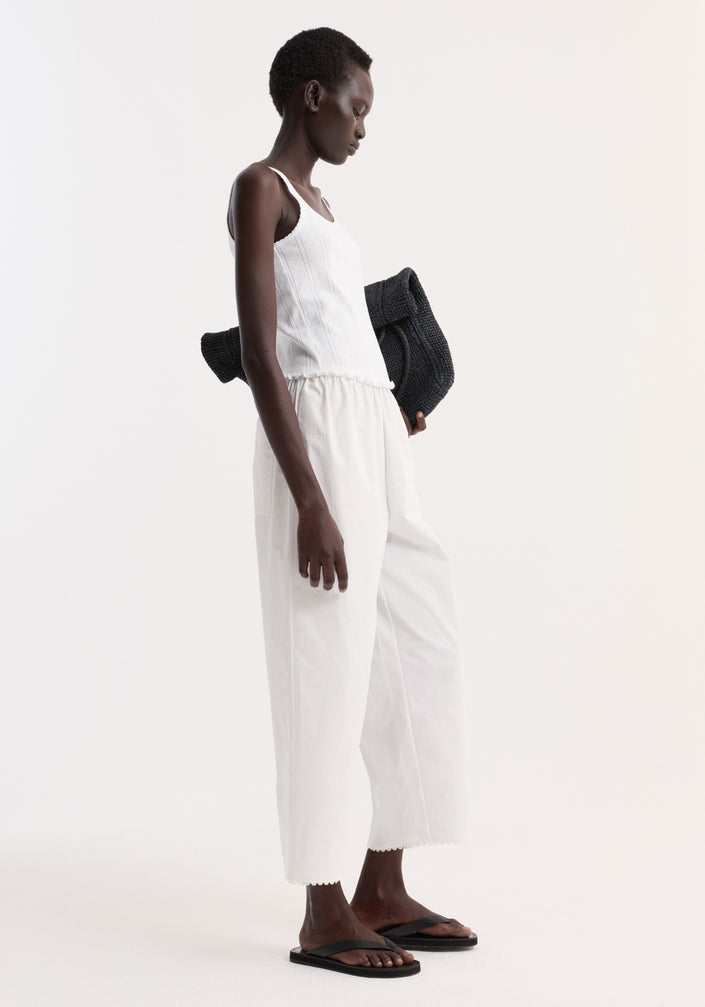 Model in Embroidered poplin trousers, optic white, with scalloped edge, high waist, and side pockets, paired with a ribbed tank top and black sandals.