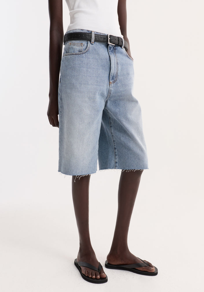 Model wearing Baggy fit denim shorts in classic blue, featuring a relaxed above-the-knee cut with raw hemline, paired with a ribbed tank top and sandals.