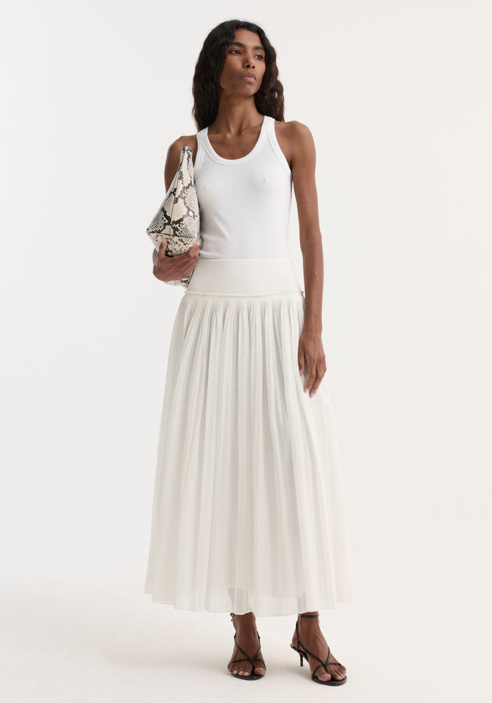 A woman models the Pleated cotton muslin skirt in optic white, featuring a fitted waistband and pleated design, paired with black sandals and a snakeskin clutch.