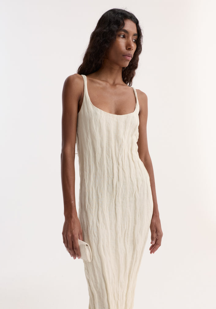 Woman wearing Textured square neck dress | cream, ankle-length, sleeveless, scooped neck. Holds matching clutch, black sandals, dark wavy hair, neutral expression.