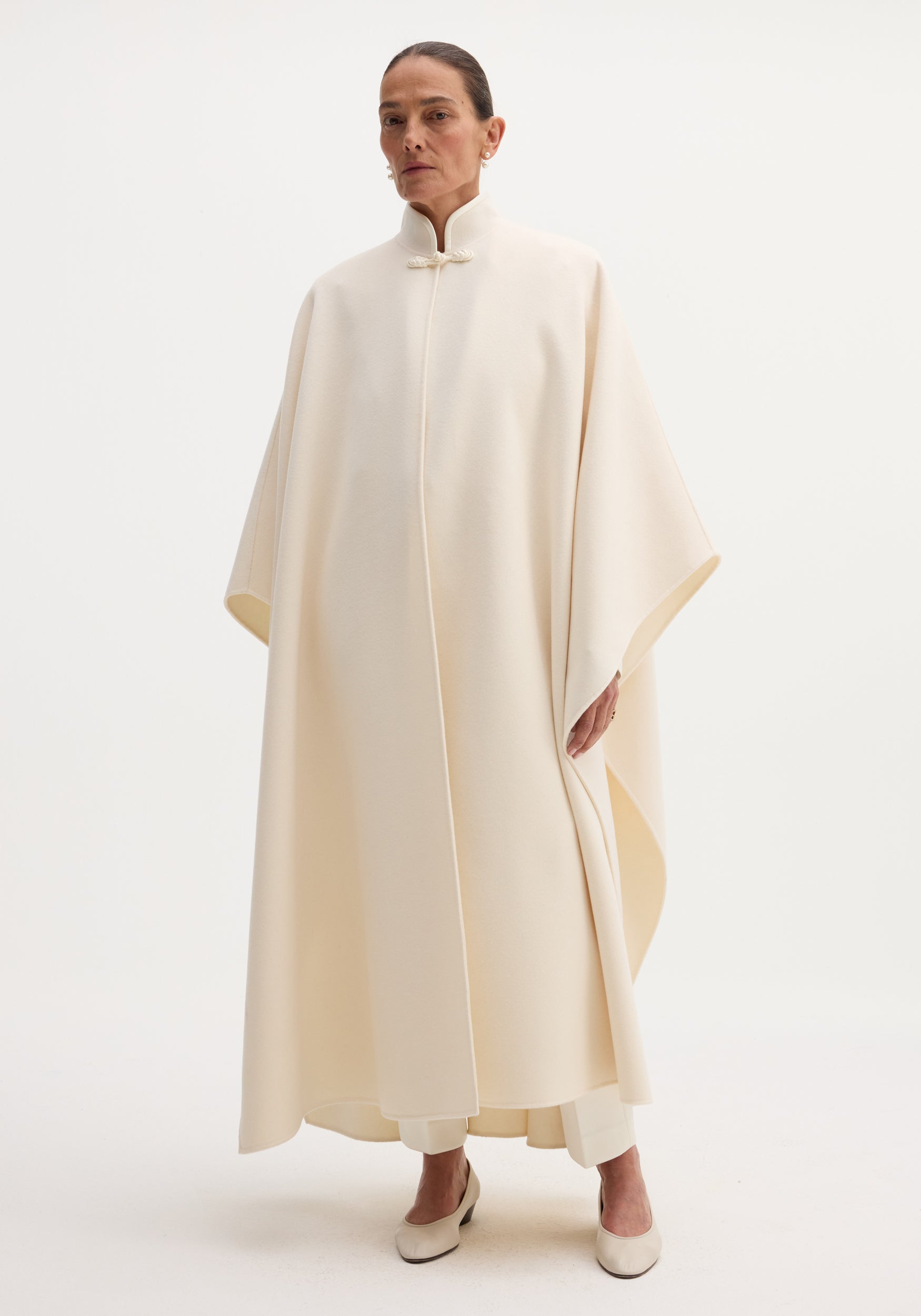 Pankou closure poncho | off white: Oversized cream cape with mandarin collar, pankou closure, light trousers, and matching low-heeled pumps, highlighting minimalist, flowing design.