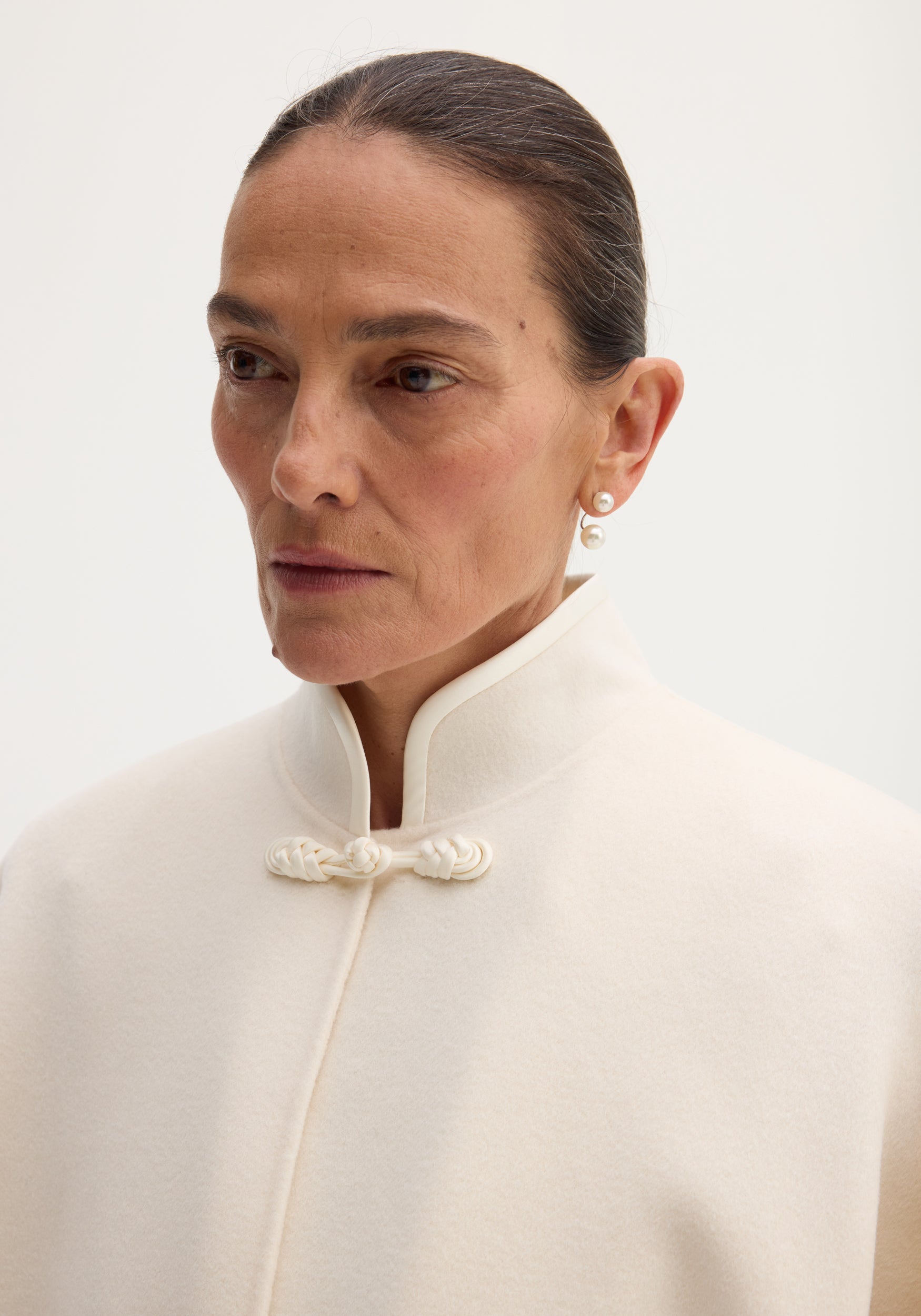 Woman wearing pearl earrings, high-collared cream coat. Showcasing Pankou closure poncho in off-white wool with stand collar, highlighting sophisticated layering.