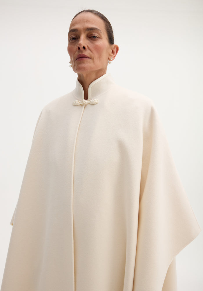 Pankou closure poncho in off-white wool, features oversized silhouette and stand collar. Modeled by a woman with pulled-back hair and pearl earrings.