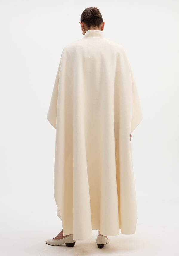Pankou closure poncho in off-white: model with tidy bun wears a full-length, oversized wool poncho, showcasing the stand collar and single pankou closure.