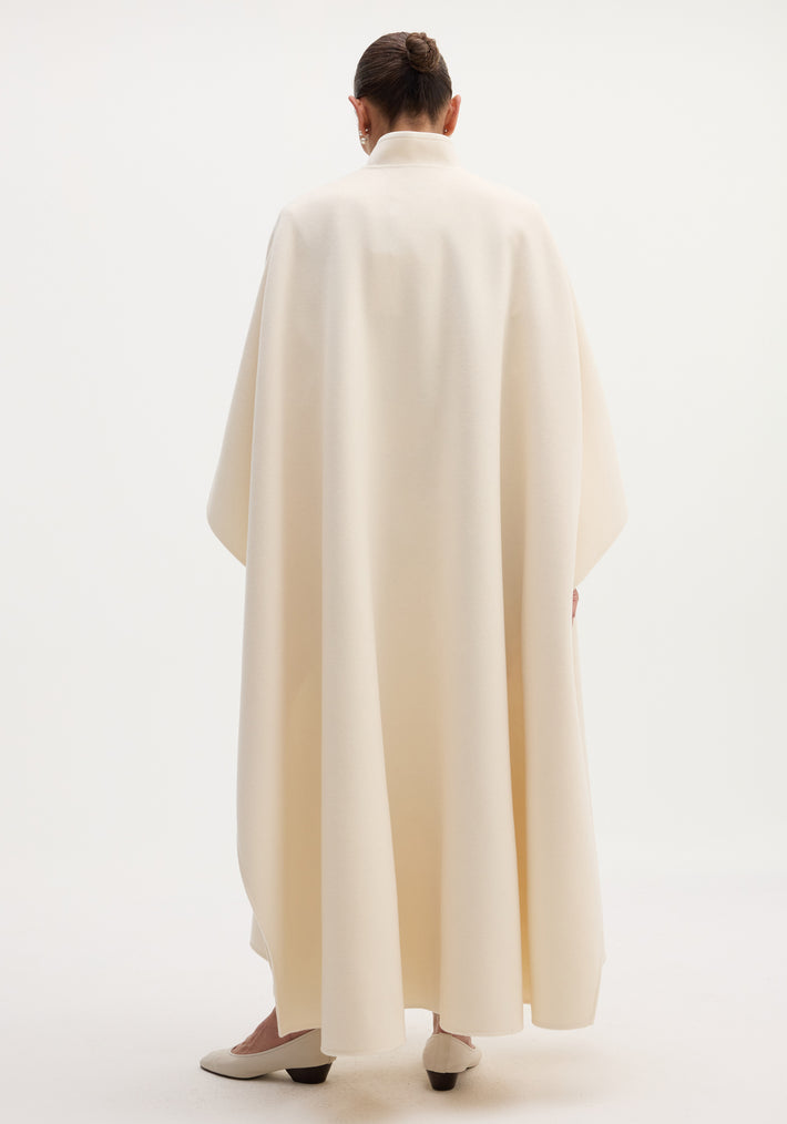 Pankou closure poncho in off-white: model with tidy bun wears a full-length, oversized wool poncho, showcasing the stand collar and single pankou closure.