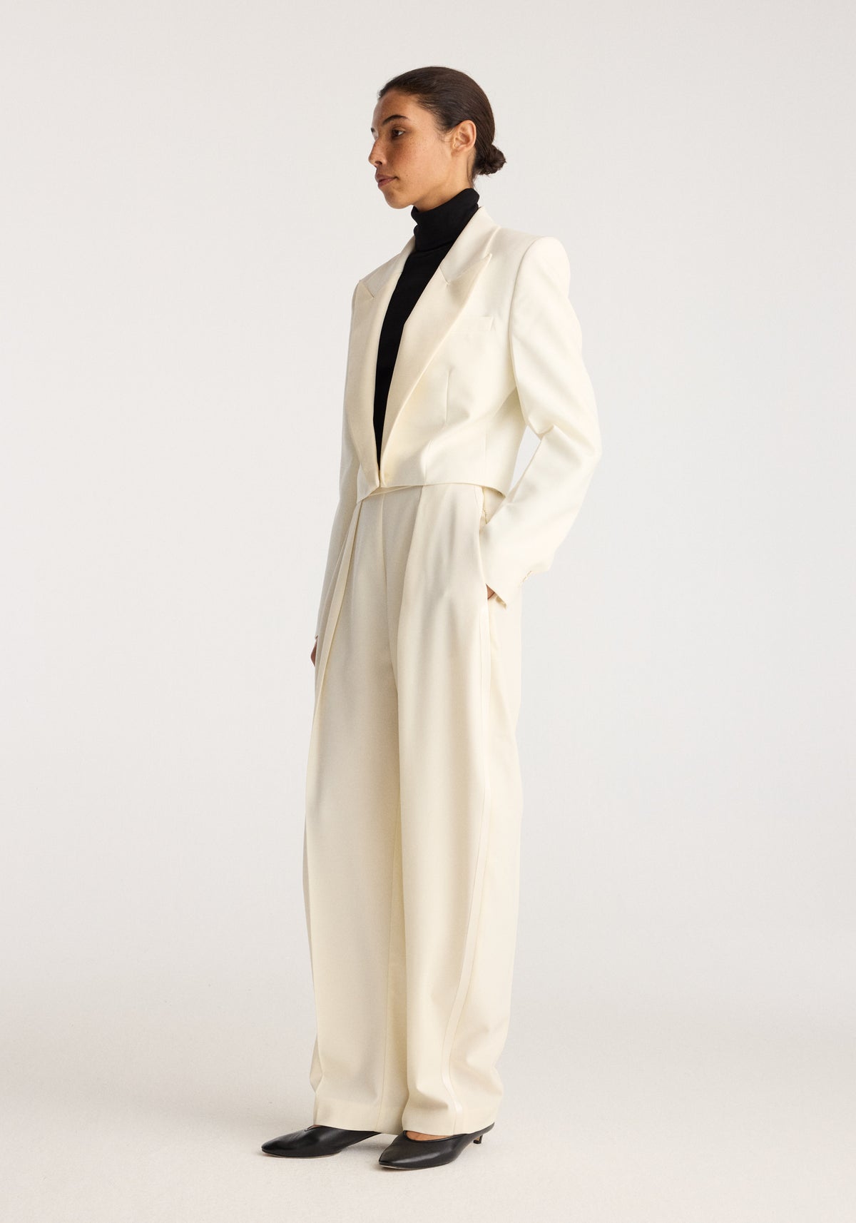 Cropped blazer | off white worn with a black turtleneck and wide leg trousers on a model in side profile against a neutral studio background