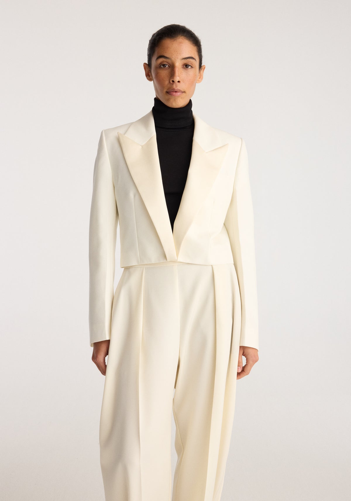 Cropped blazer | off white worn by a model with a black turtleneck, featuring peaked lapels, long sleeves, and a fitted cropped silhouette