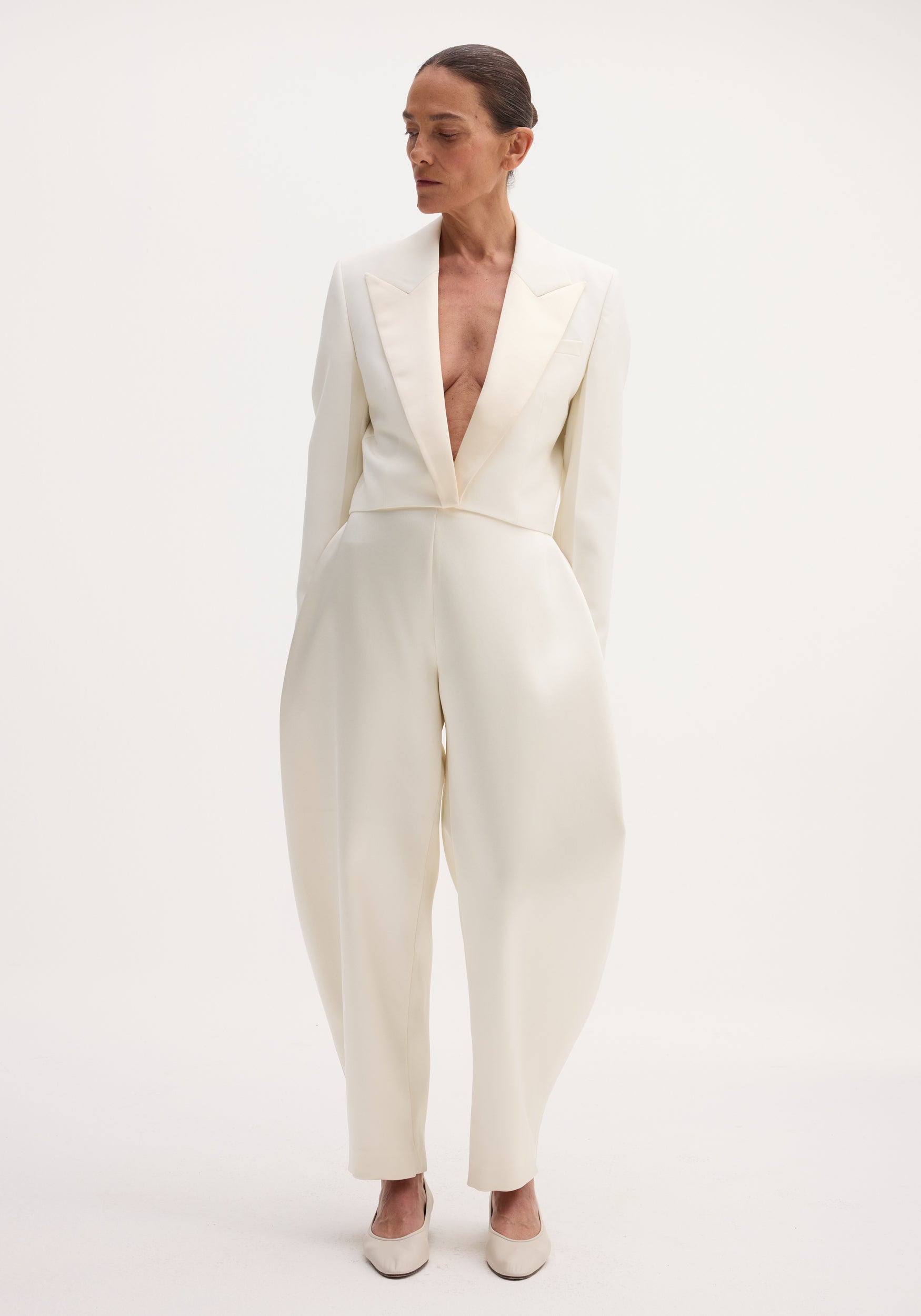 Model wearing Sculptural satin trousers in ivory, featuring a high waist and bold balloon legs, paired with a notched-lapel jacket and flats.