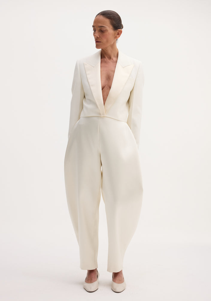 Model wearing Sculptural satin trousers in ivory, featuring a high waist and bold balloon legs, paired with a notched-lapel jacket and flats.
