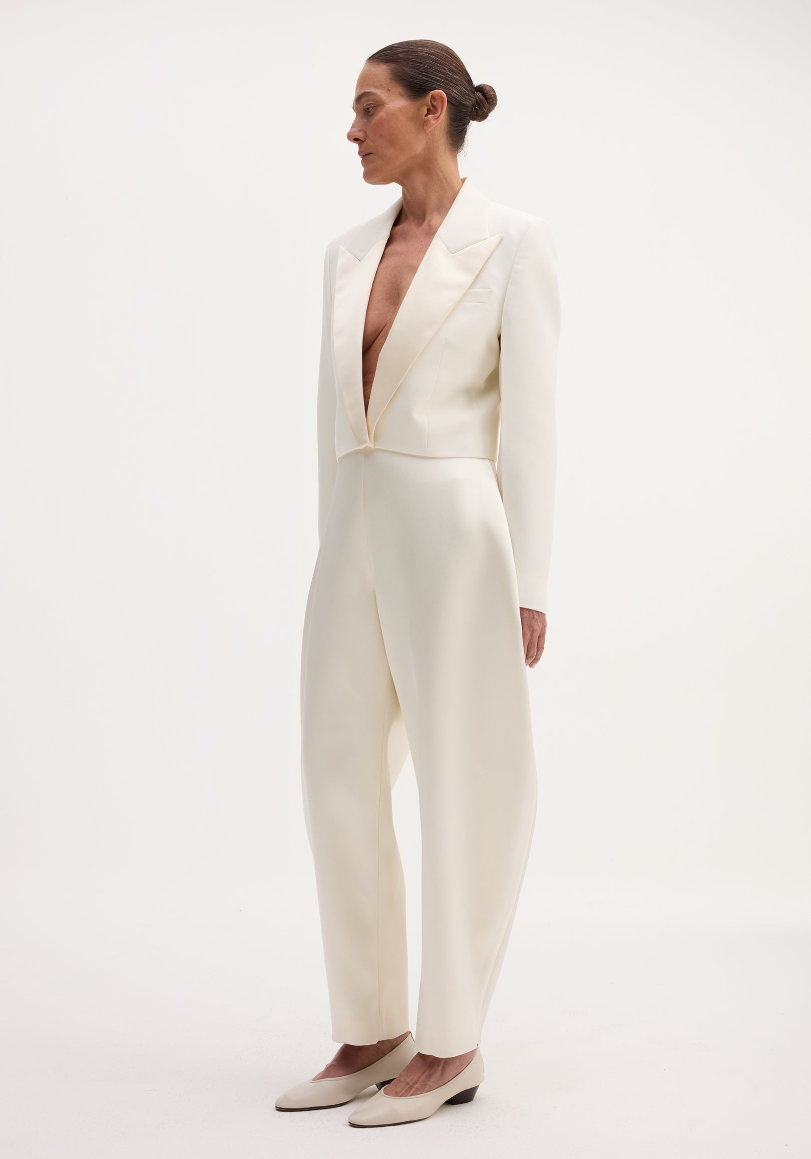 Model in a Cropped blazer | off white with satin peak lapels, high-waisted trousers, and low-heeled shoes, showcasing a sleek, monochromatic look.