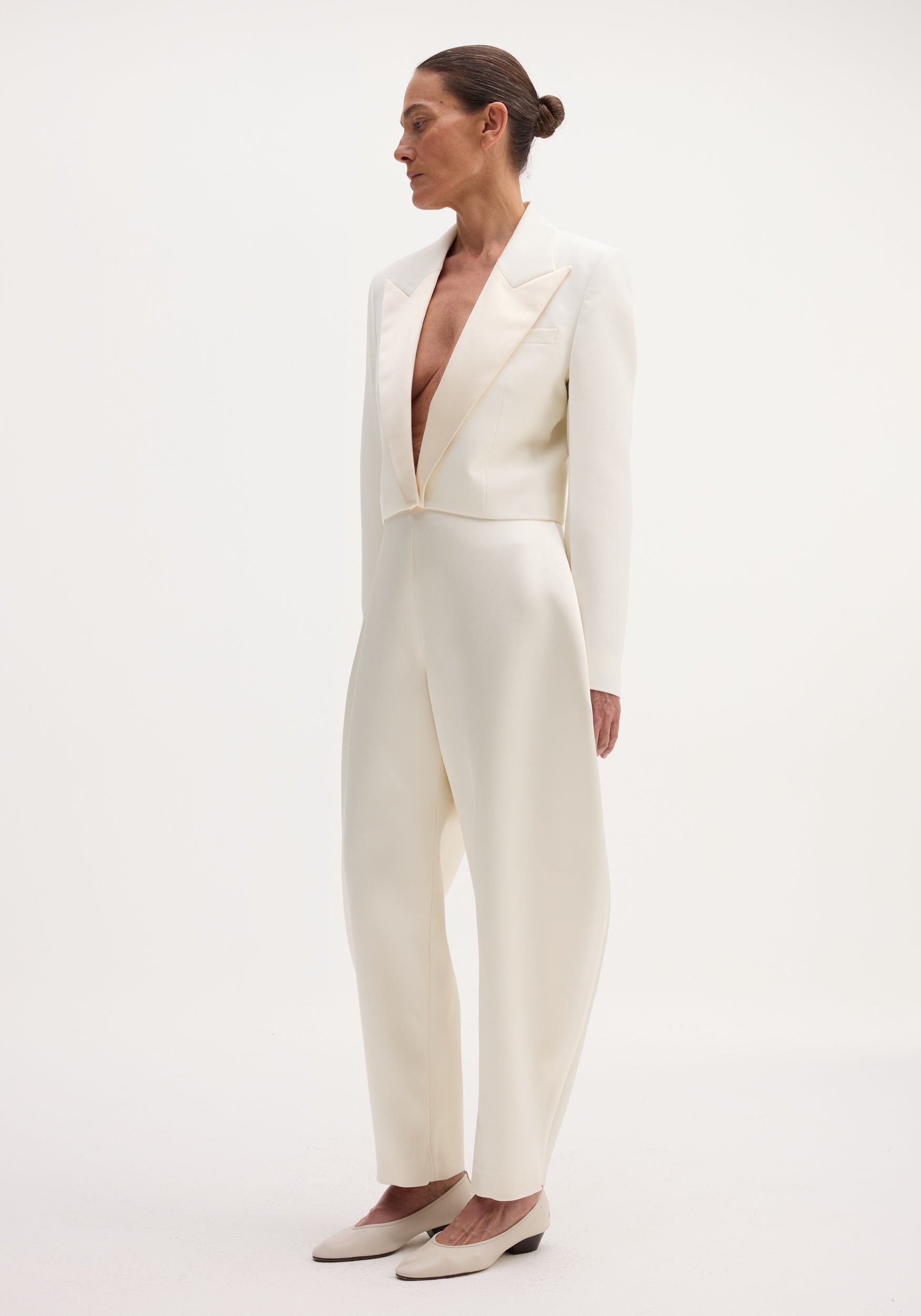 Model in a Cropped blazer | off white with satin peak lapels, high-waisted trousers, and low-heeled shoes, showcasing a sleek, monochromatic look.