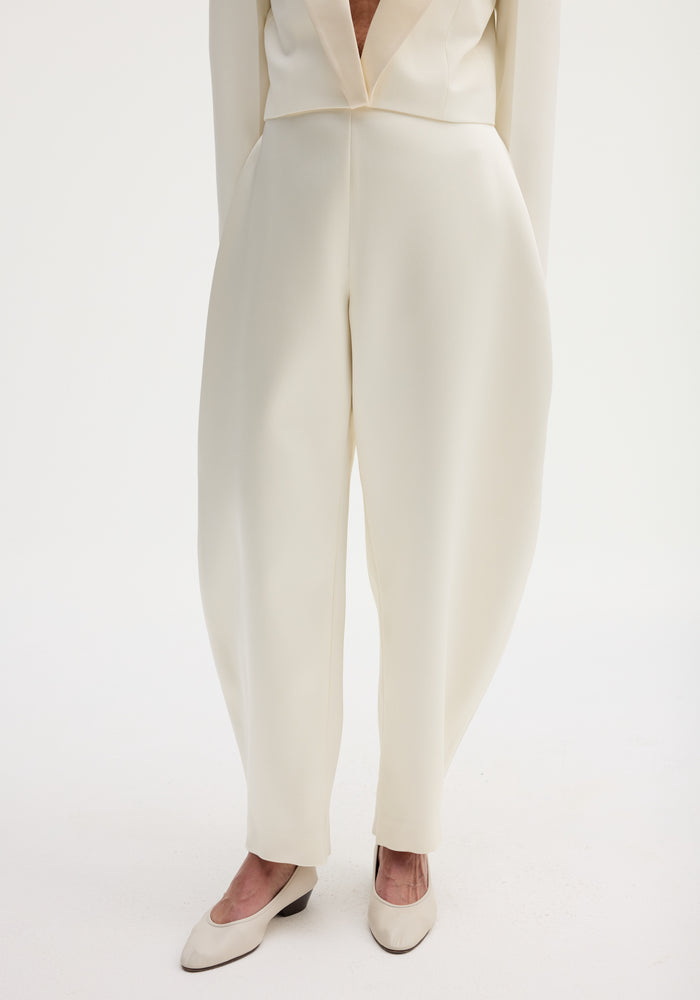 Lower half of a person in sculptural satin trousers with a high waist and balloon leg, paired with matching low-heeled pumps, showcasing a modern silhouette.