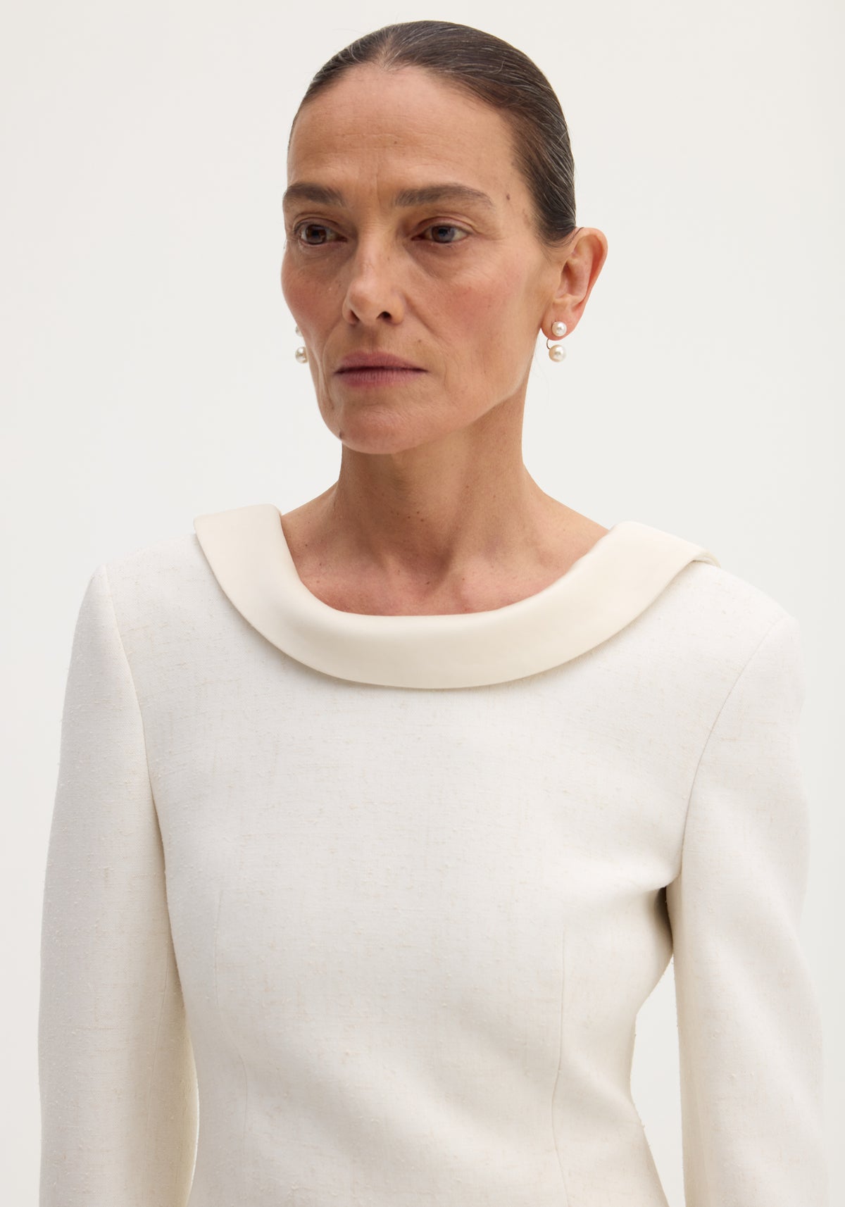 Woman wearing a reversed blazer in winter white, featuring a softly draped neckline, drop pearl earrings, and structured fabric with button closures and welt pockets.
