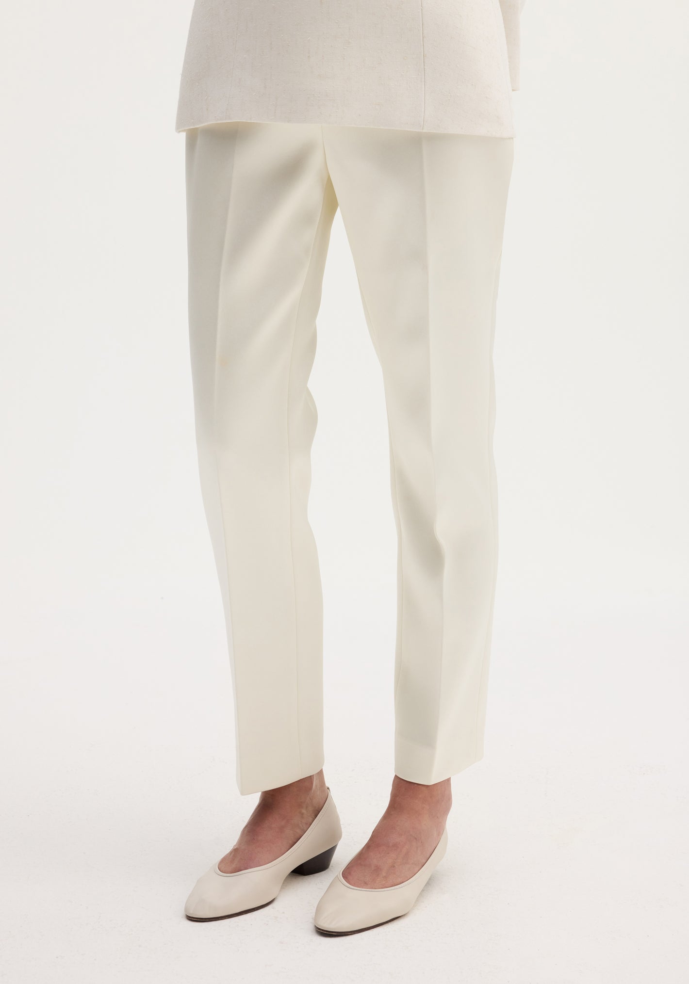 Cigarette leg satin trousers in ivory, featuring a high-rise fit and sharp center crease, paired with matching block-heeled pumps on a plain background.