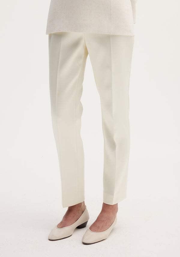 Cigarette leg satin trousers in ivory, featuring a high-rise fit and sharp center crease, paired with matching block-heeled pumps on a plain background.