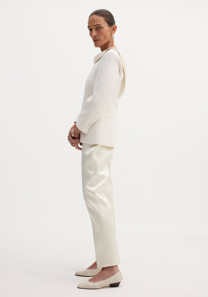 Woman in profile wearing ivory cigarette leg satin trousers, featuring pin-tuck detailing, ankle length, high-rise fit, paired with a tailored jacket and leather flats.