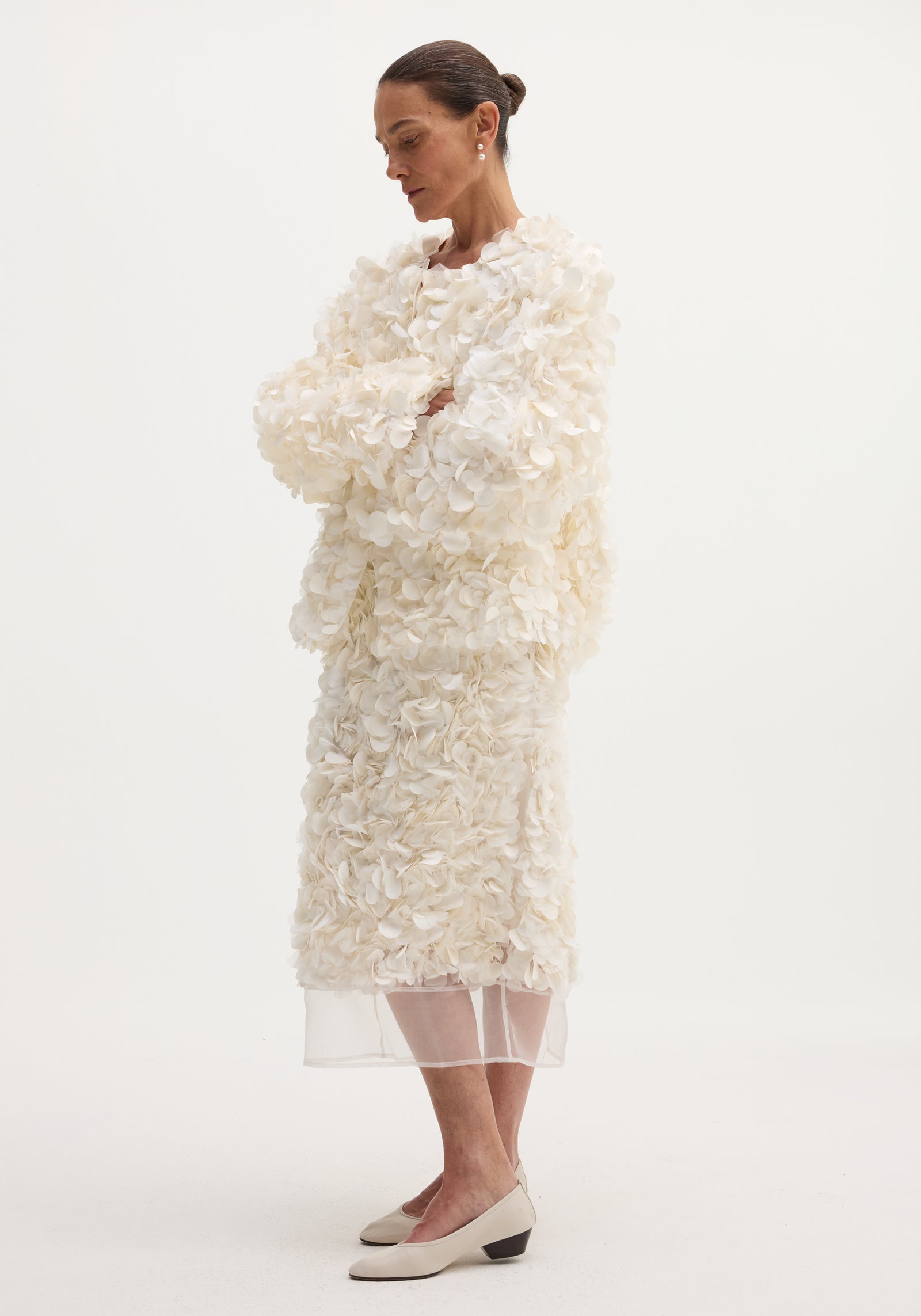 Handmade embellished jacket in ivory with petal-shaped fabric appliqués, worn by a woman with folded arms. Features a hook-and-eye closure at the neckline.