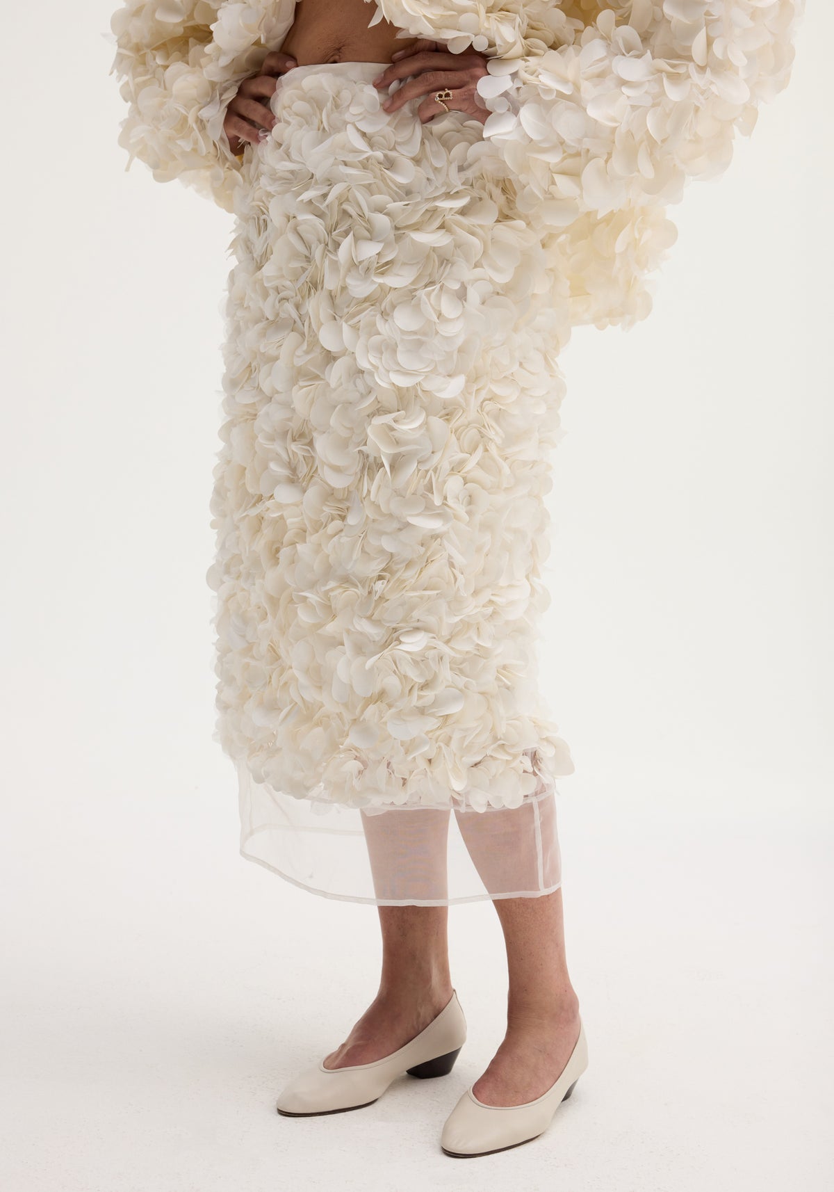 Handmade embellished skirt in ivory, featuring a voluminous knee-length design with delicate fabric appliqués and a refined silk organza hem, modeled with cream low-heeled pumps.