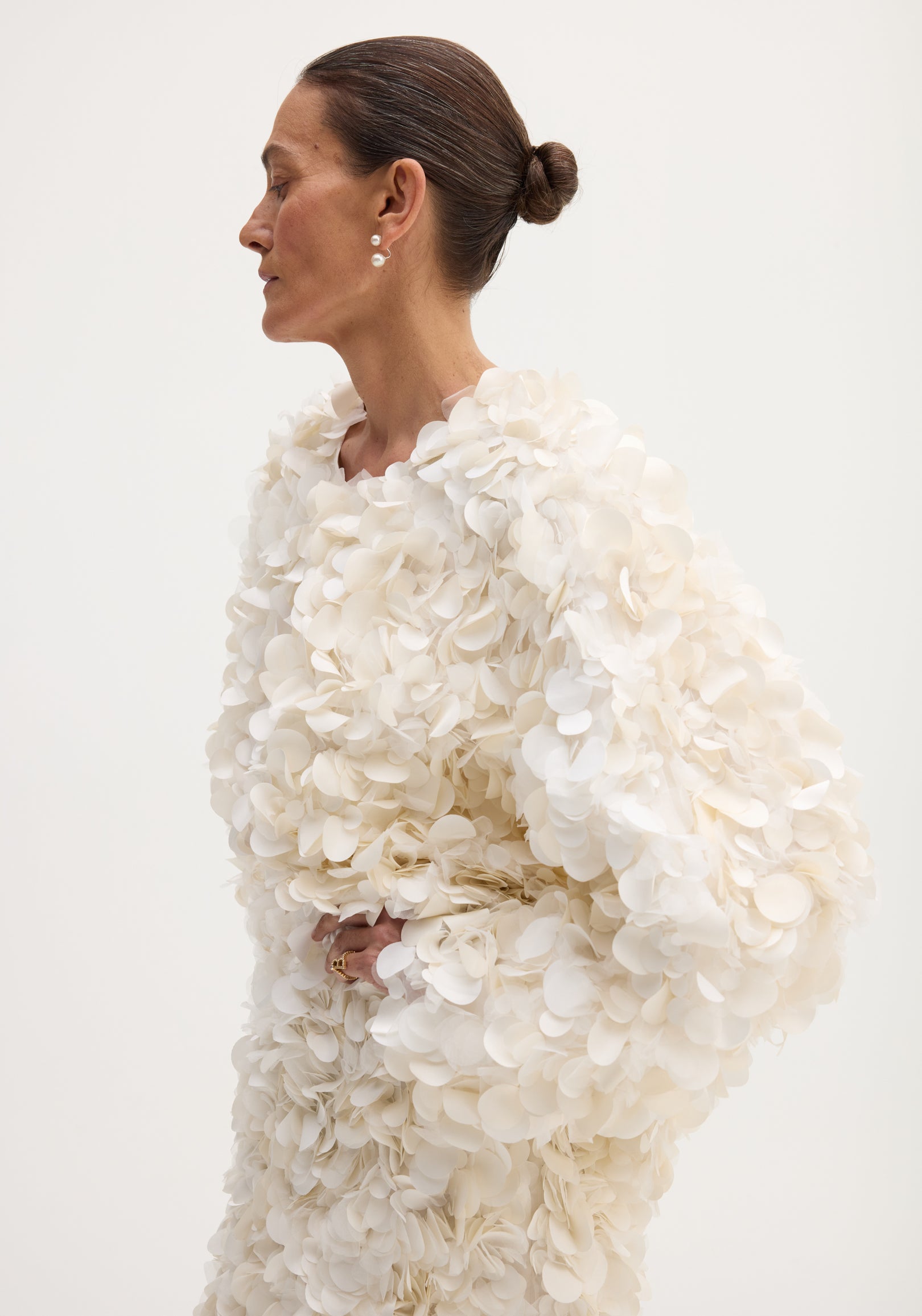 Side-profile of a woman in a Handmade embellished jacket | ivory, featuring petal-shaped appliqués and pearl earrings, with a hook-and-eye closure at the neckline.