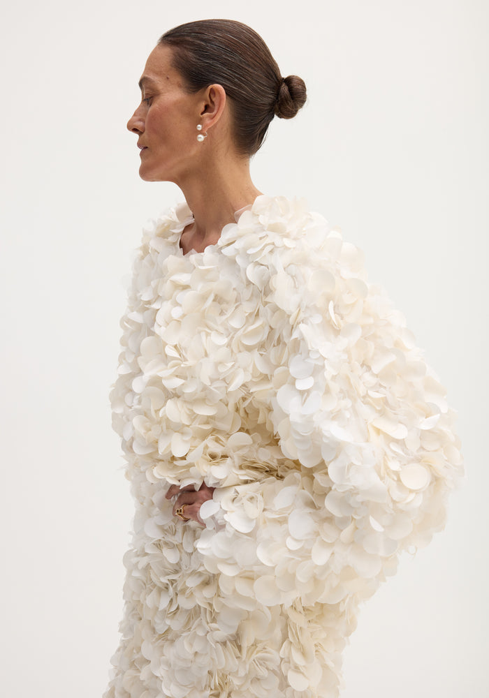 Side-profile of a woman in a Handmade embellished jacket | ivory, featuring petal-shaped appliqués and pearl earrings, with a hook-and-eye closure at the neckline.
