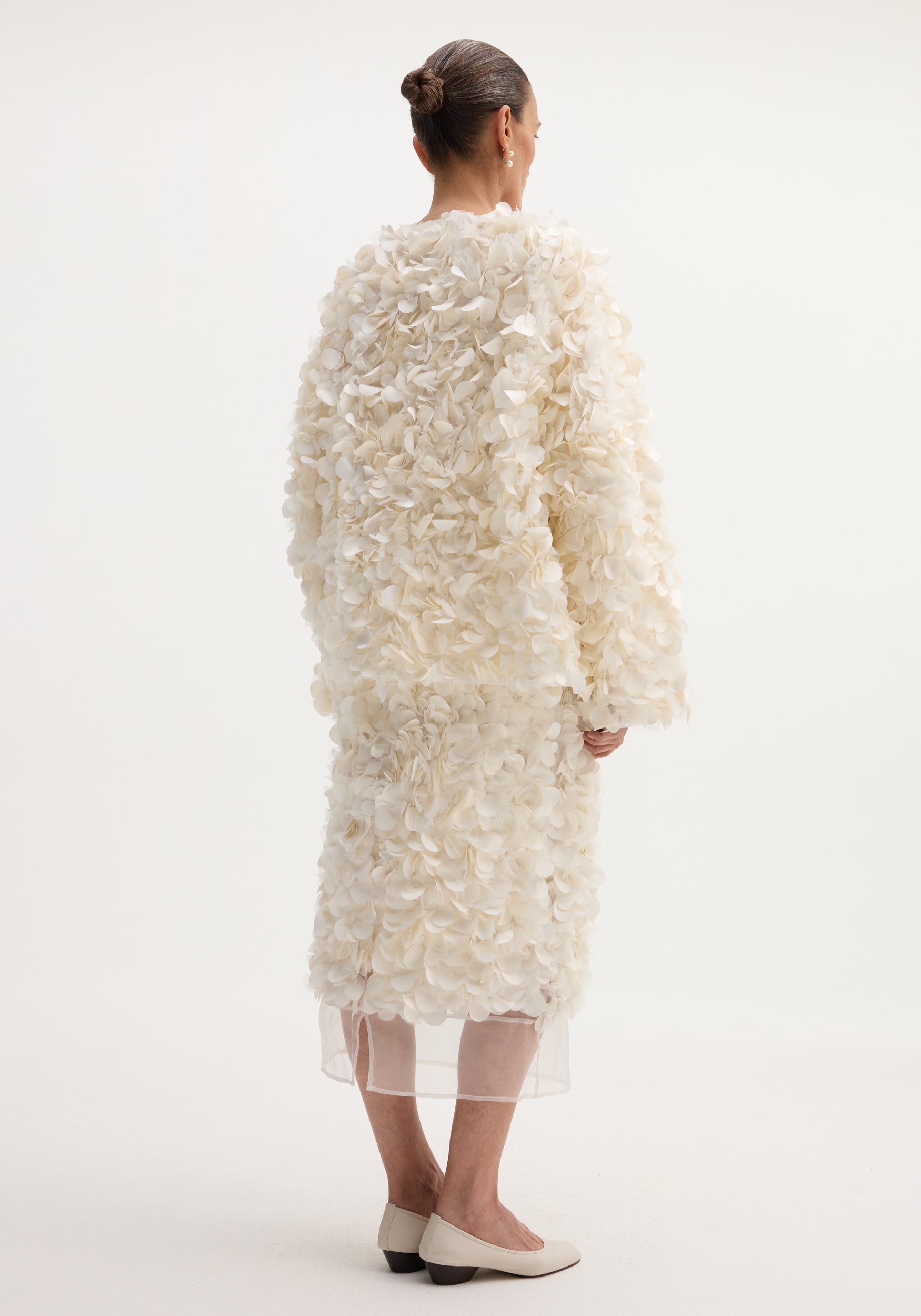 Back view of a woman wearing a Handmade embellished jacket in ivory, featuring petal-like appliqués, over a matching skirt, showcasing the jacket's voluminous design.