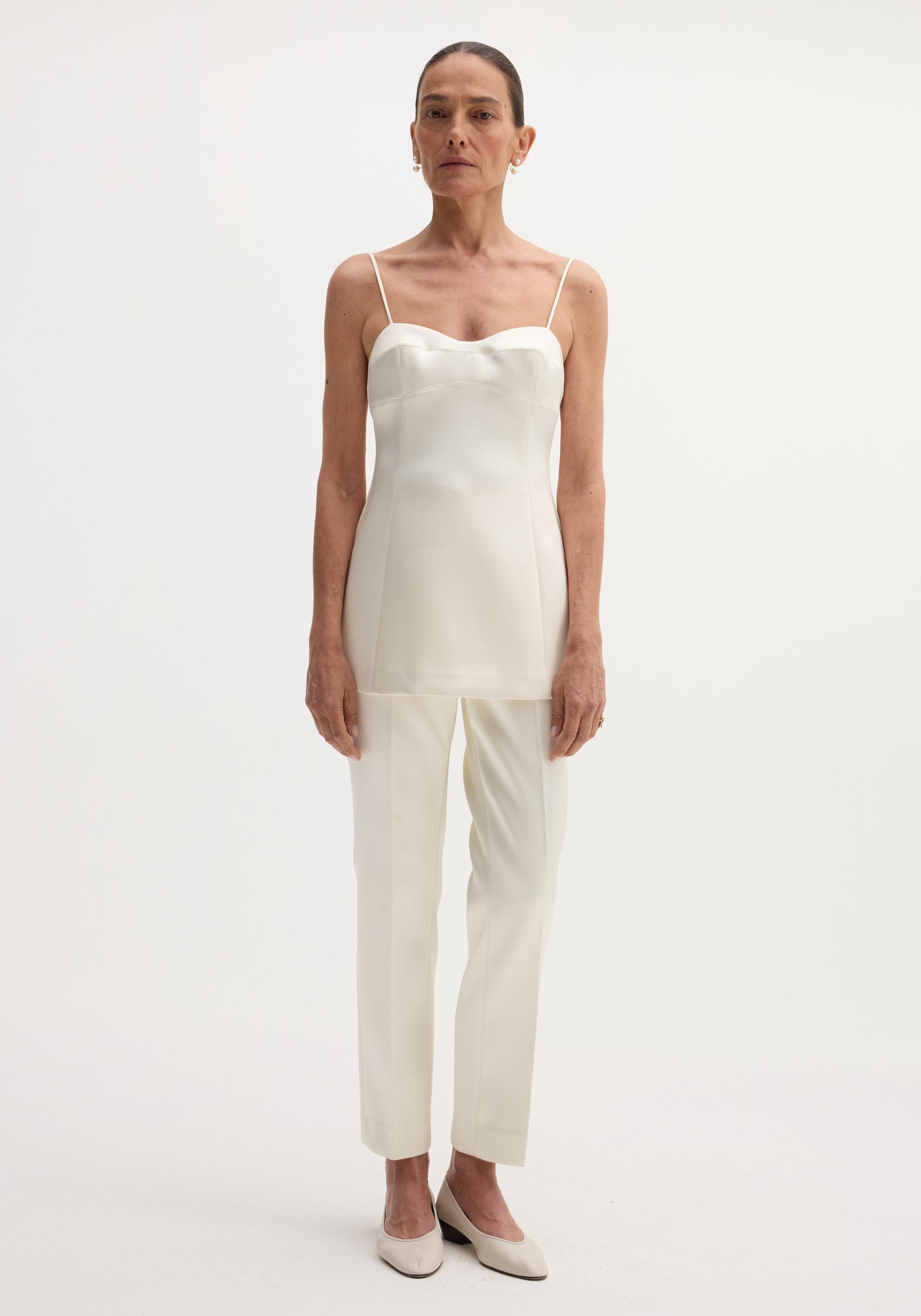 Model wears Satin bustier top | ivory, featuring a fitted bustier-style design, sweetheart neckline, and adjustable back straps, paired with matching trousers and pearl earrings.
