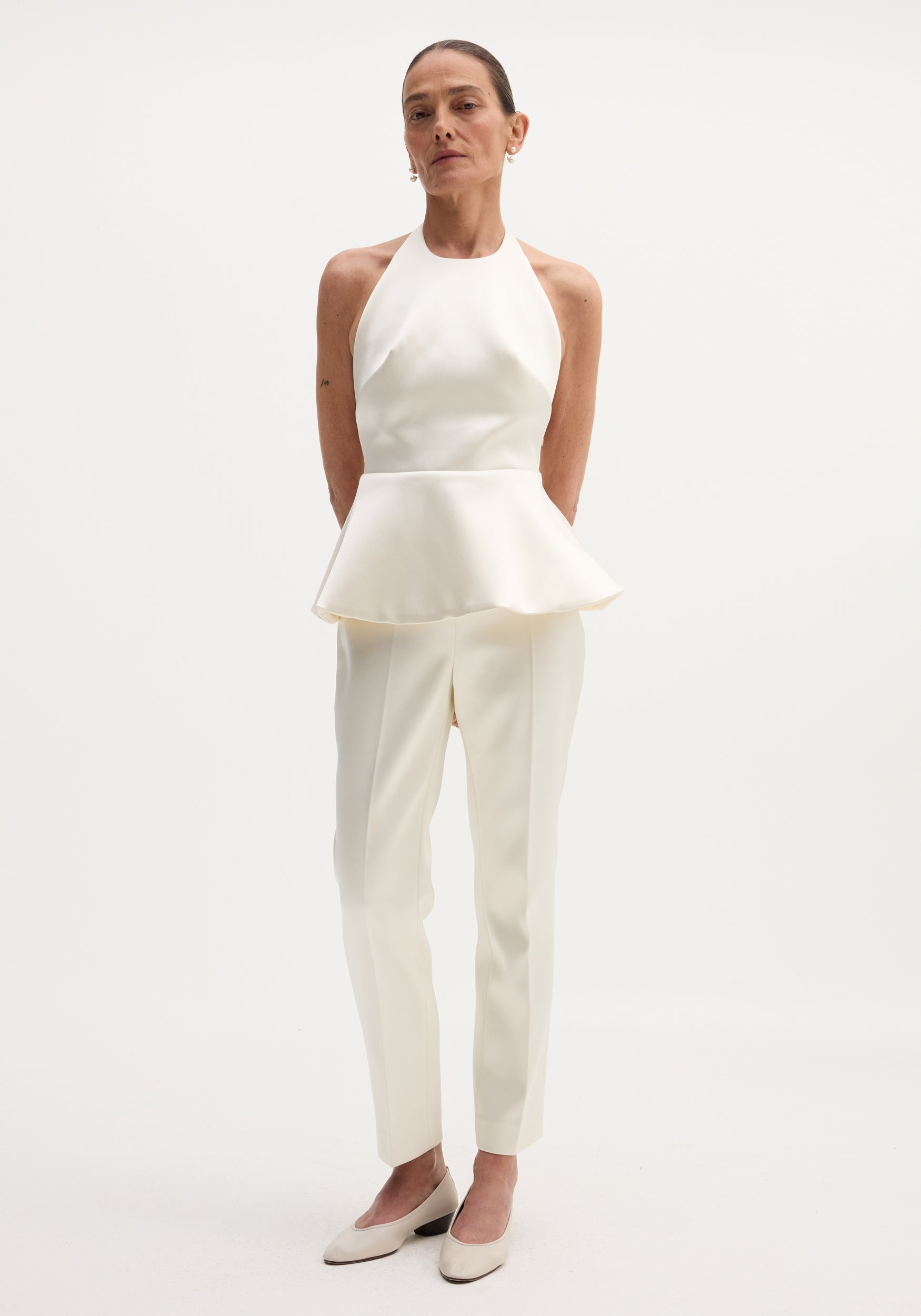 Model wearing Sculptural satin halter top with a fitted bodice and peplum, paired with matching tailored trousers and low-heeled pumps.